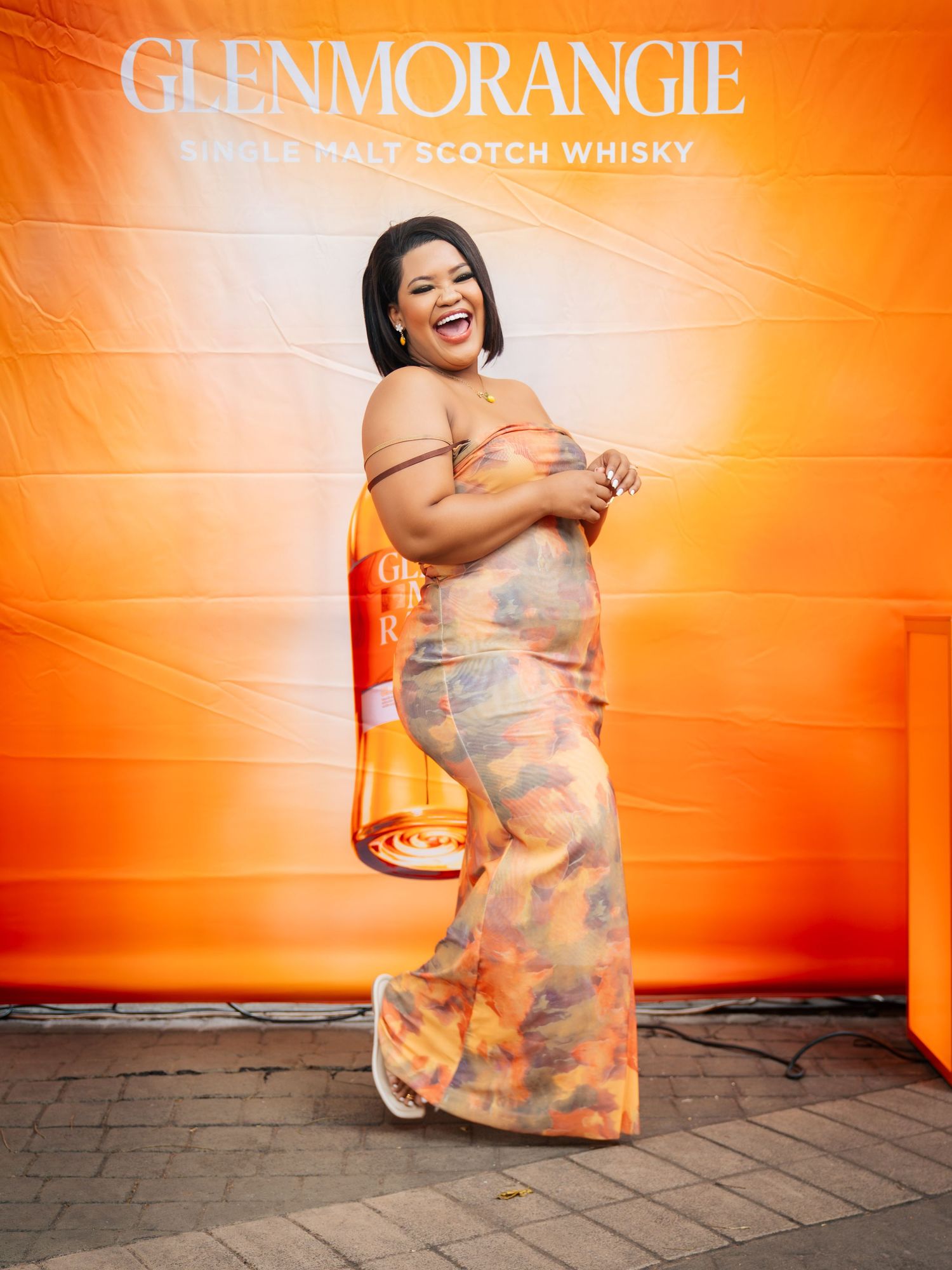 Woman in colorful maxi dress laughing at Glenmorangie Single Malt Scotch Whisky event backdrop.