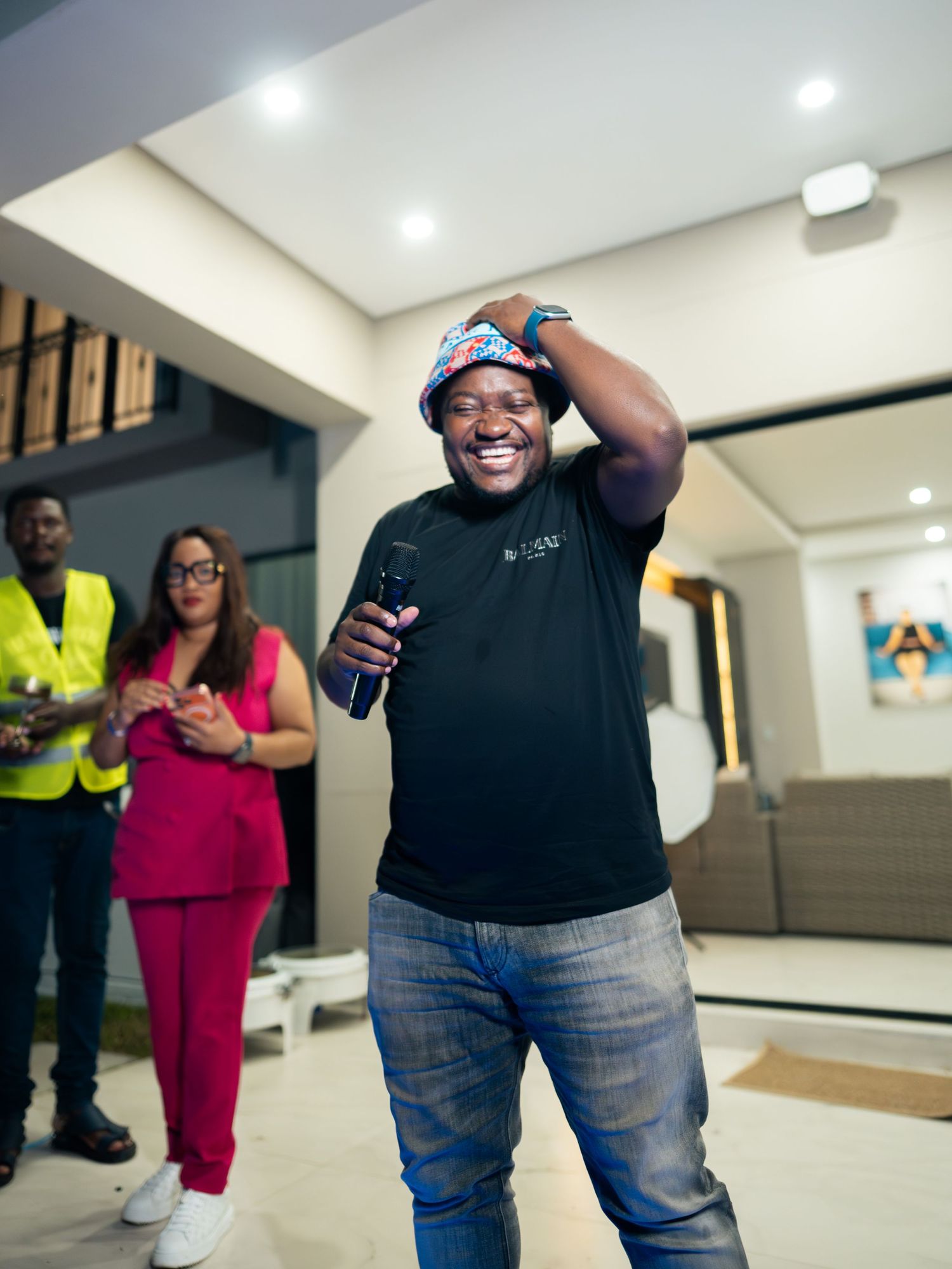 A joyful man in a black shirt and bucket hat laughs while holding a microphone at an indoor gathering.