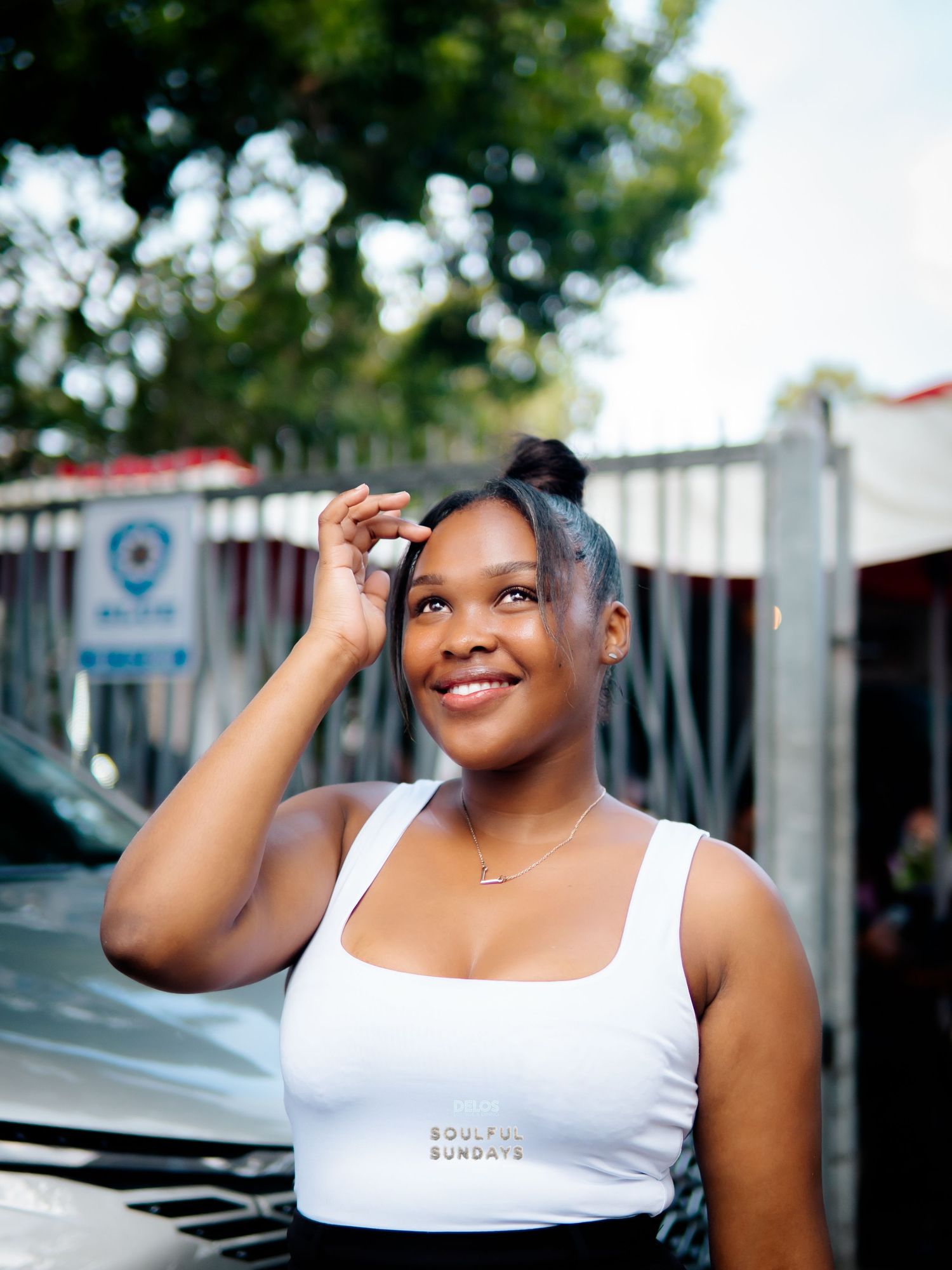 Young woman in white crop top smiling outdoors near a car, with trees and urban background.