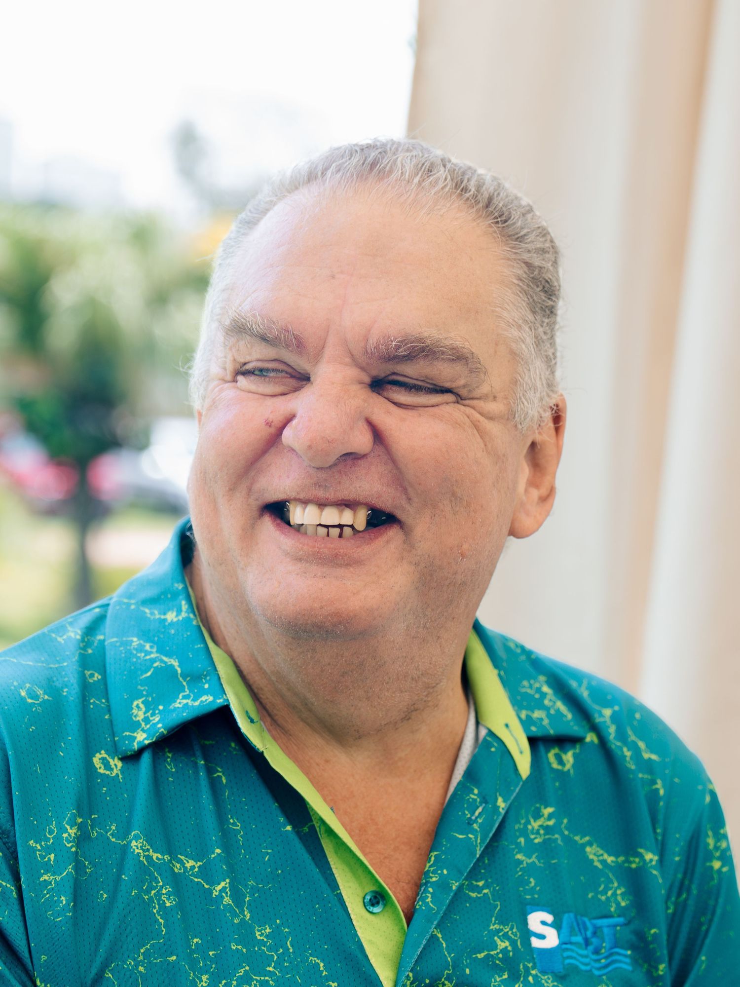 Smiling middle-aged man in a teal and green patterned shirt laughing outdoors in bright sunlight.
