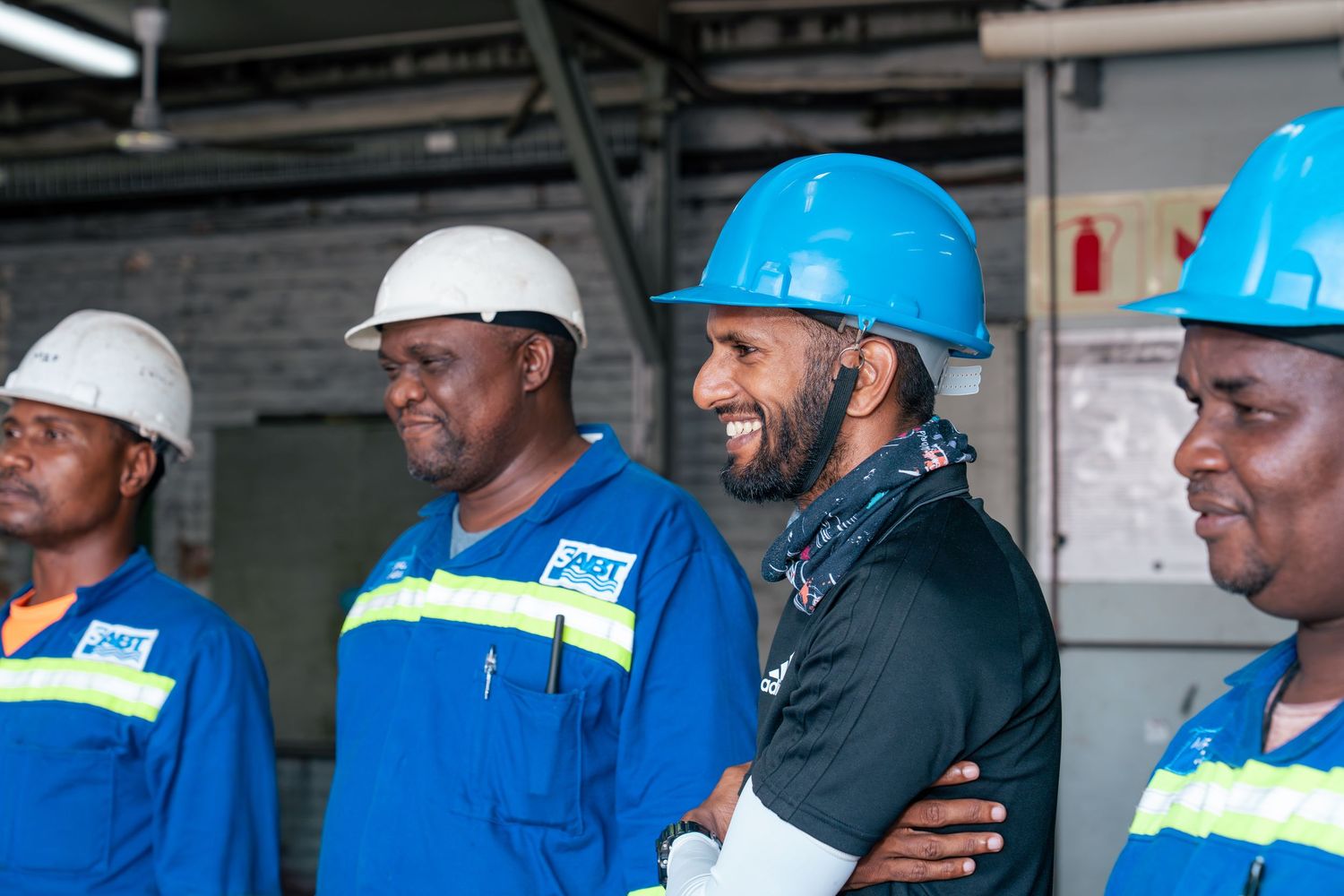 Four workers in blue uniforms and hard hats smile together at an industrial facility.