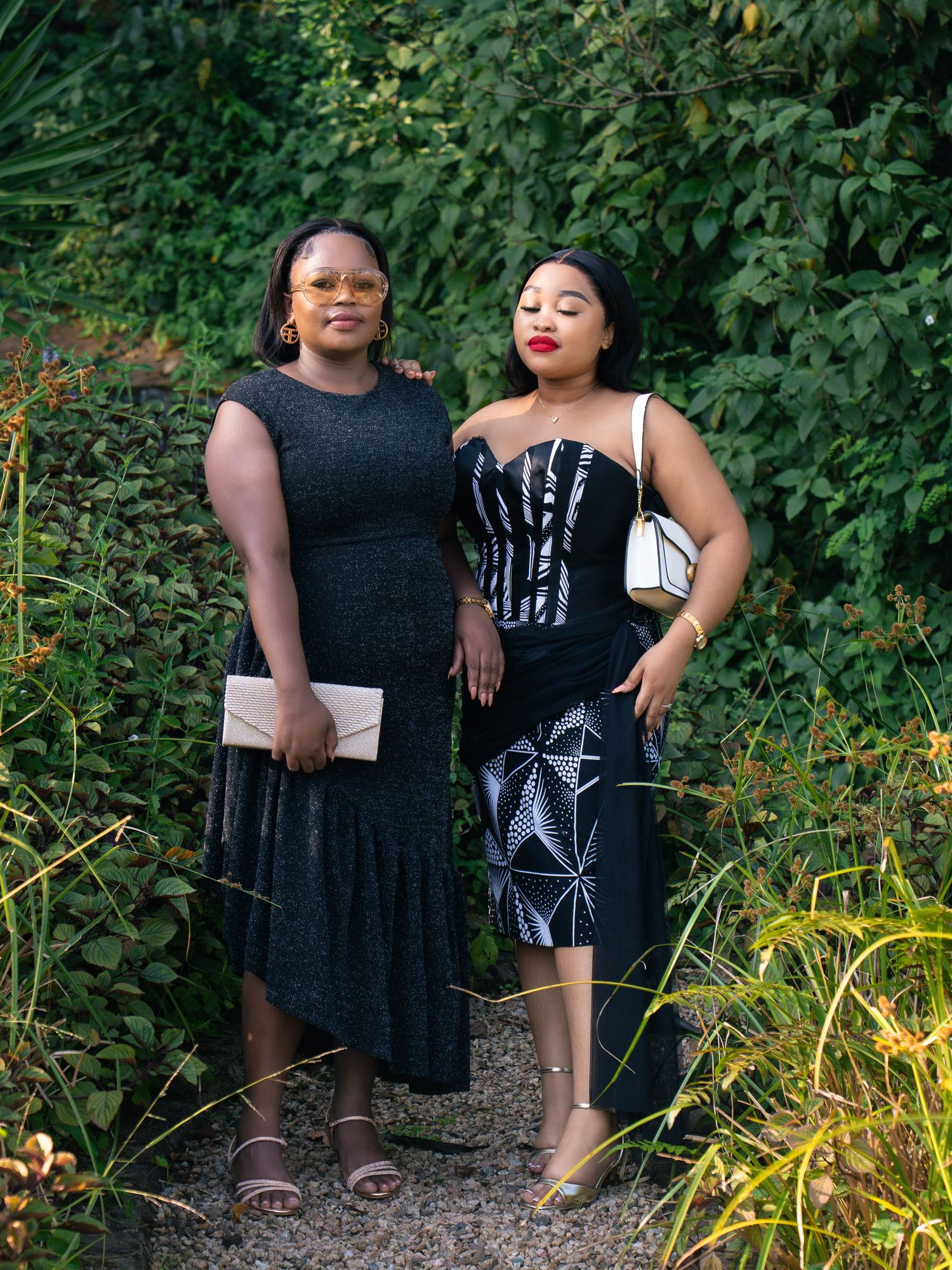 Two elegant women in black formal attire pose together outdoors among lush green foliage.