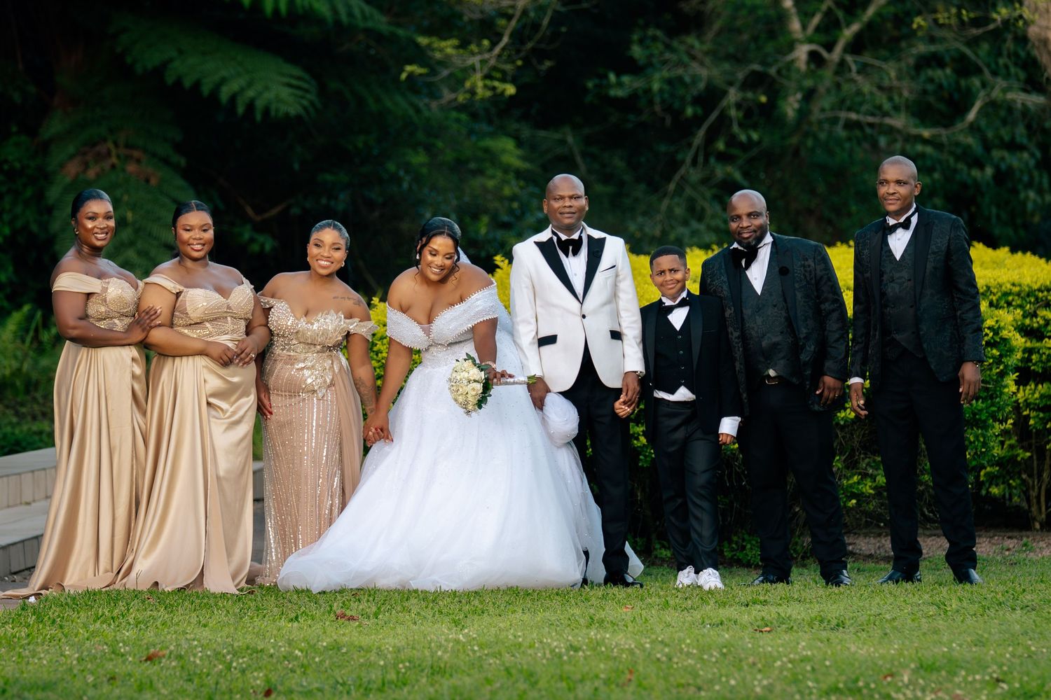 Wedding party poses outdoors, bride in white gown, groom in tuxedo, bridesmaids in gold dresses, groomsmen in dark suits.
