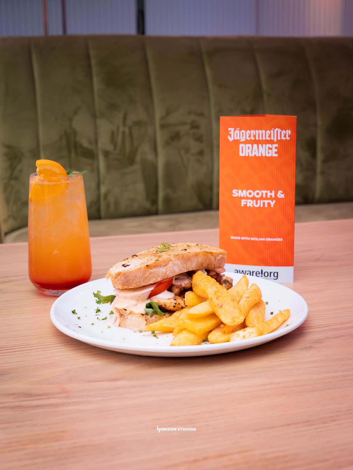 Jagermeister Orange cocktail served with a chicken sandwich and golden fries on a white plate at a restaurant.
