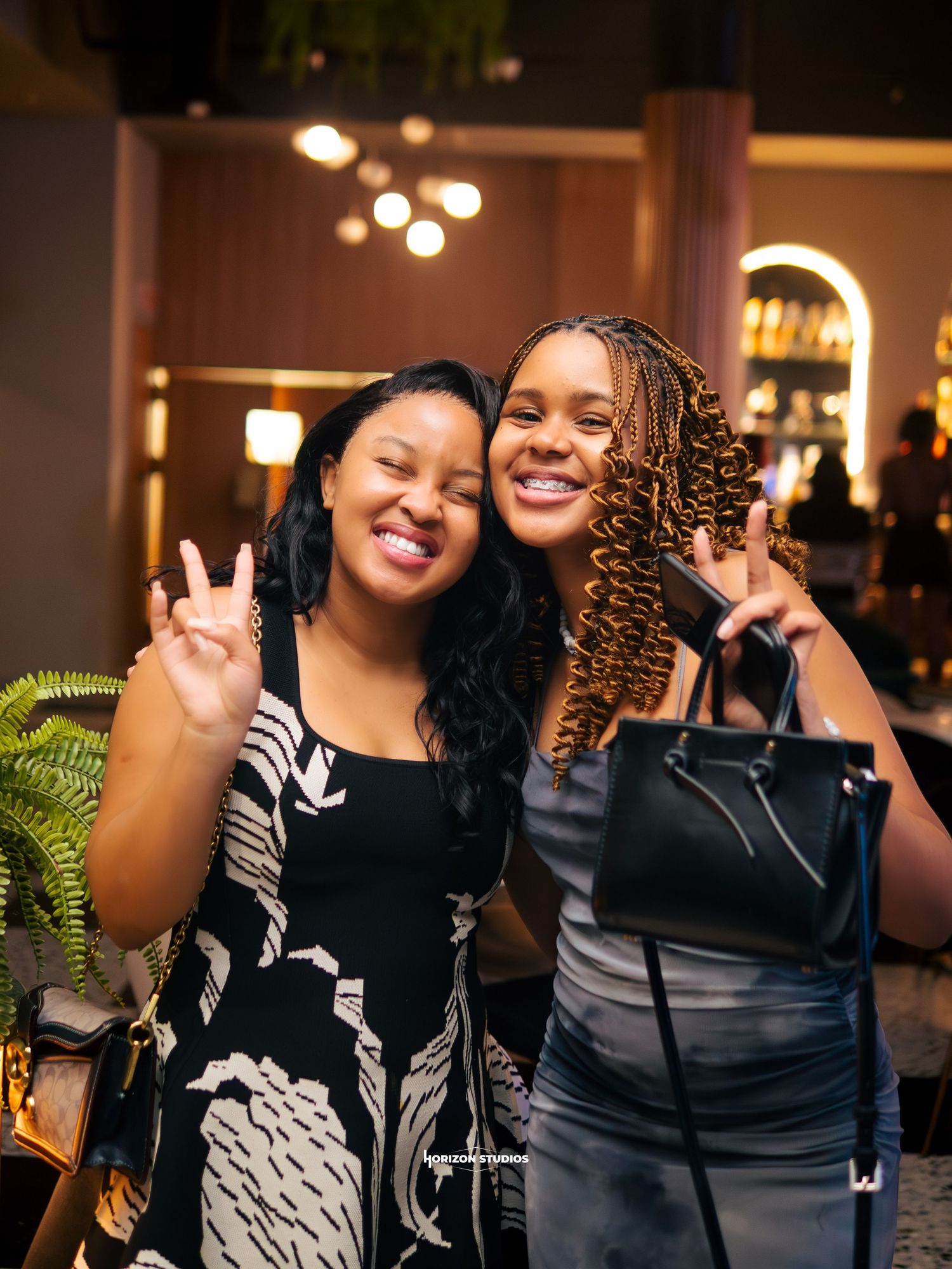 Two smiling young Black women pose making peace signs at an upscale restaurant or bar.