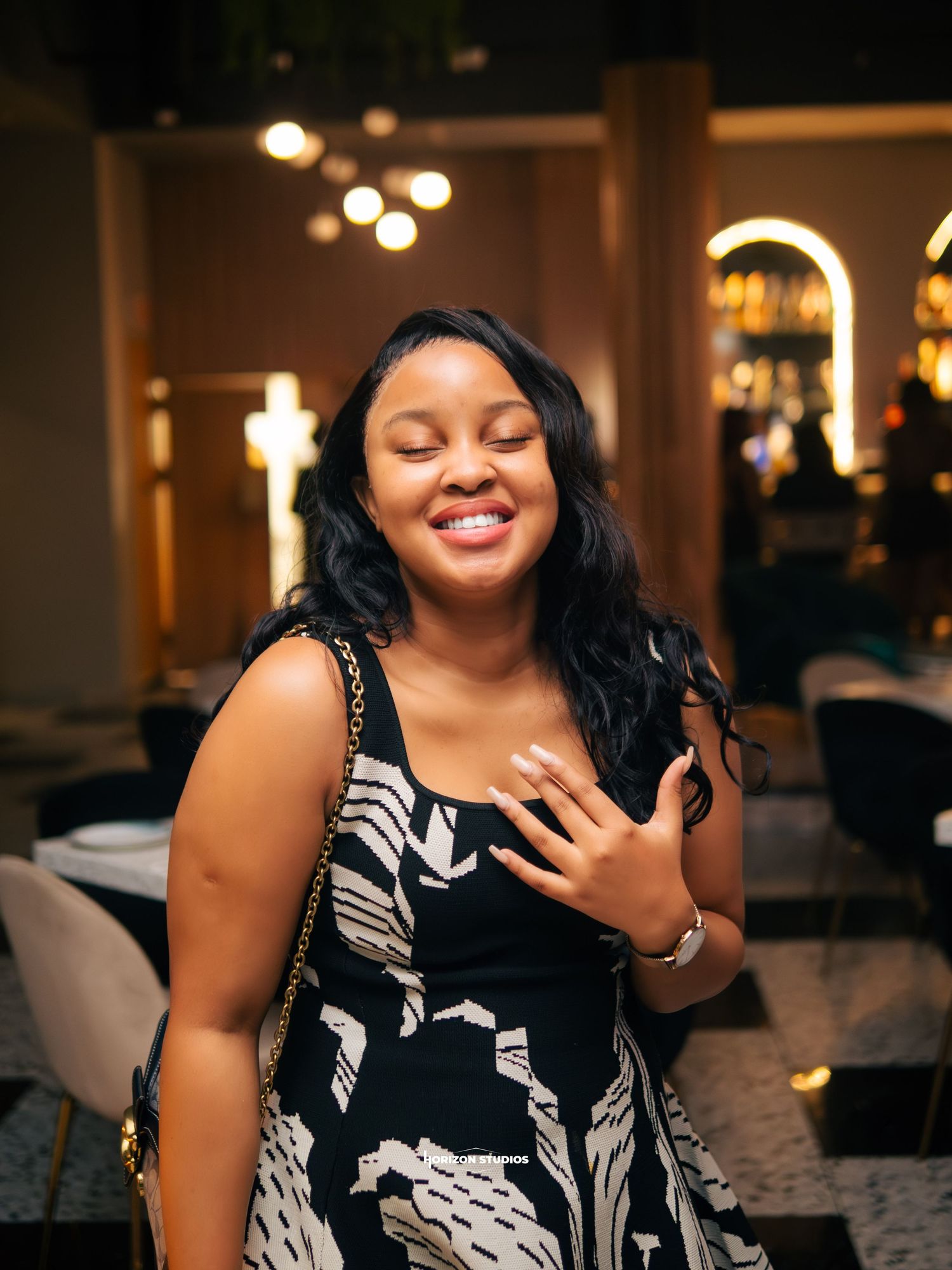Smiling young woman in black and white floral dress with crossbody bag at elegant restaurant.