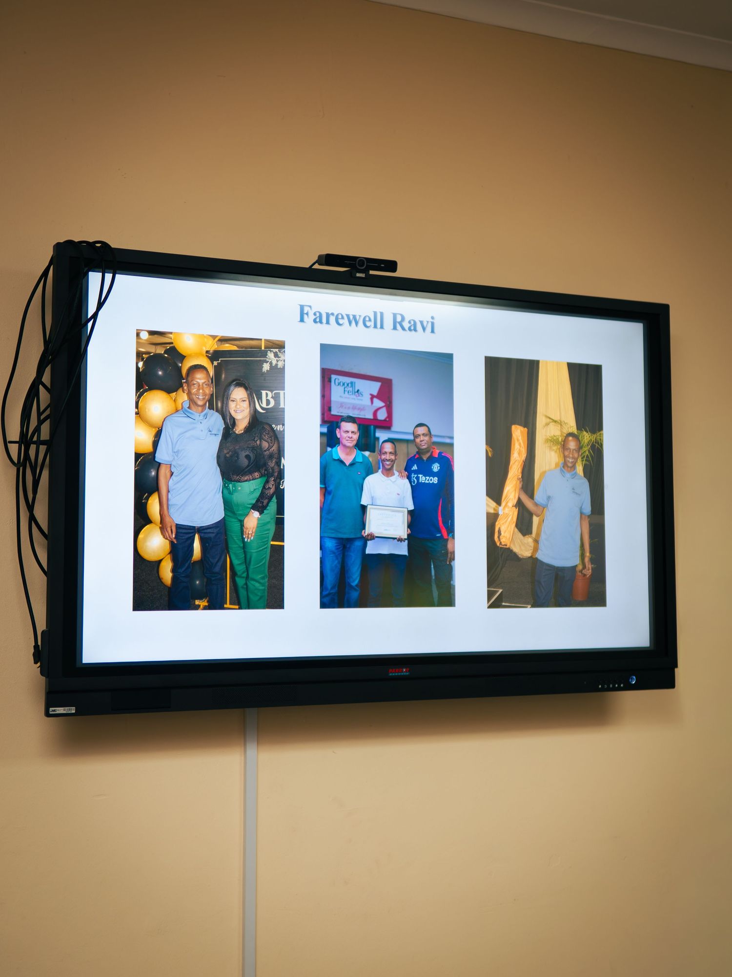 A wall-mounted TV displays a Farewell Ravi slideshow with three celebratory photos of colleagues at a goodbye party.