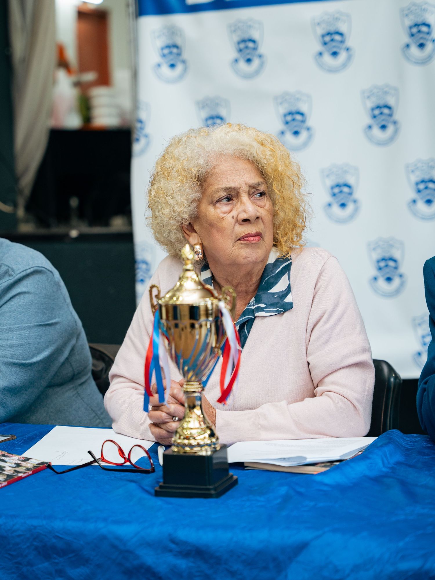 Elderly woman with curly blonde hair holds a gold trophy at a formal table with a blue banner backdrop.