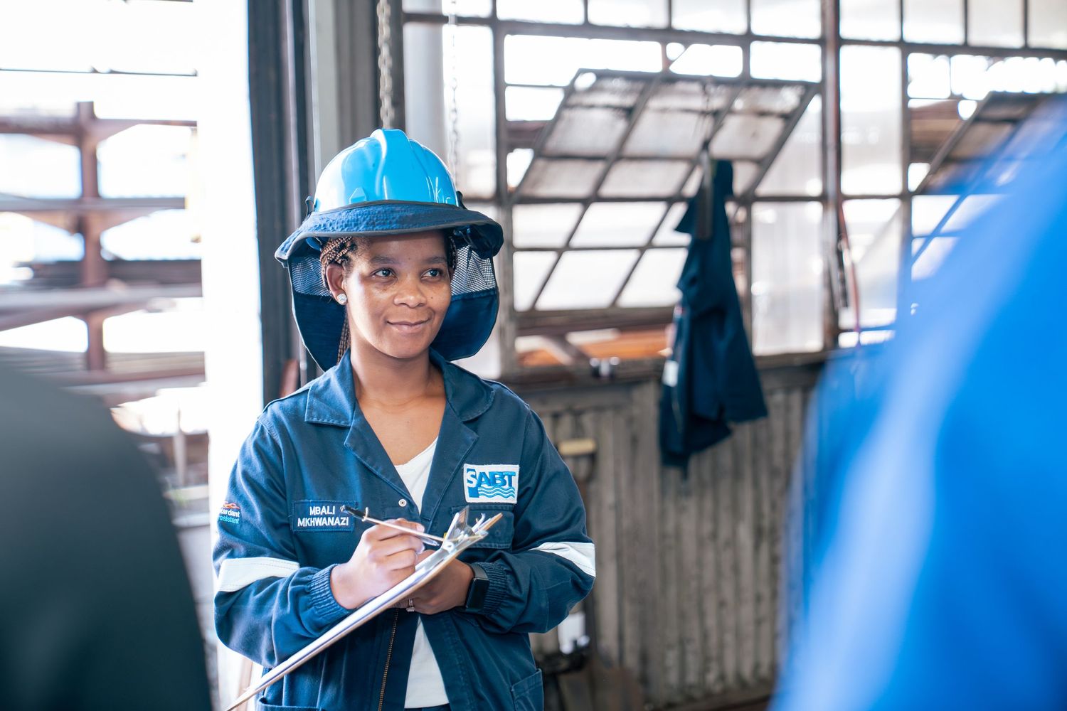 Female engineer in blue hard hat and coveralls holds clipboard in industrial facility.