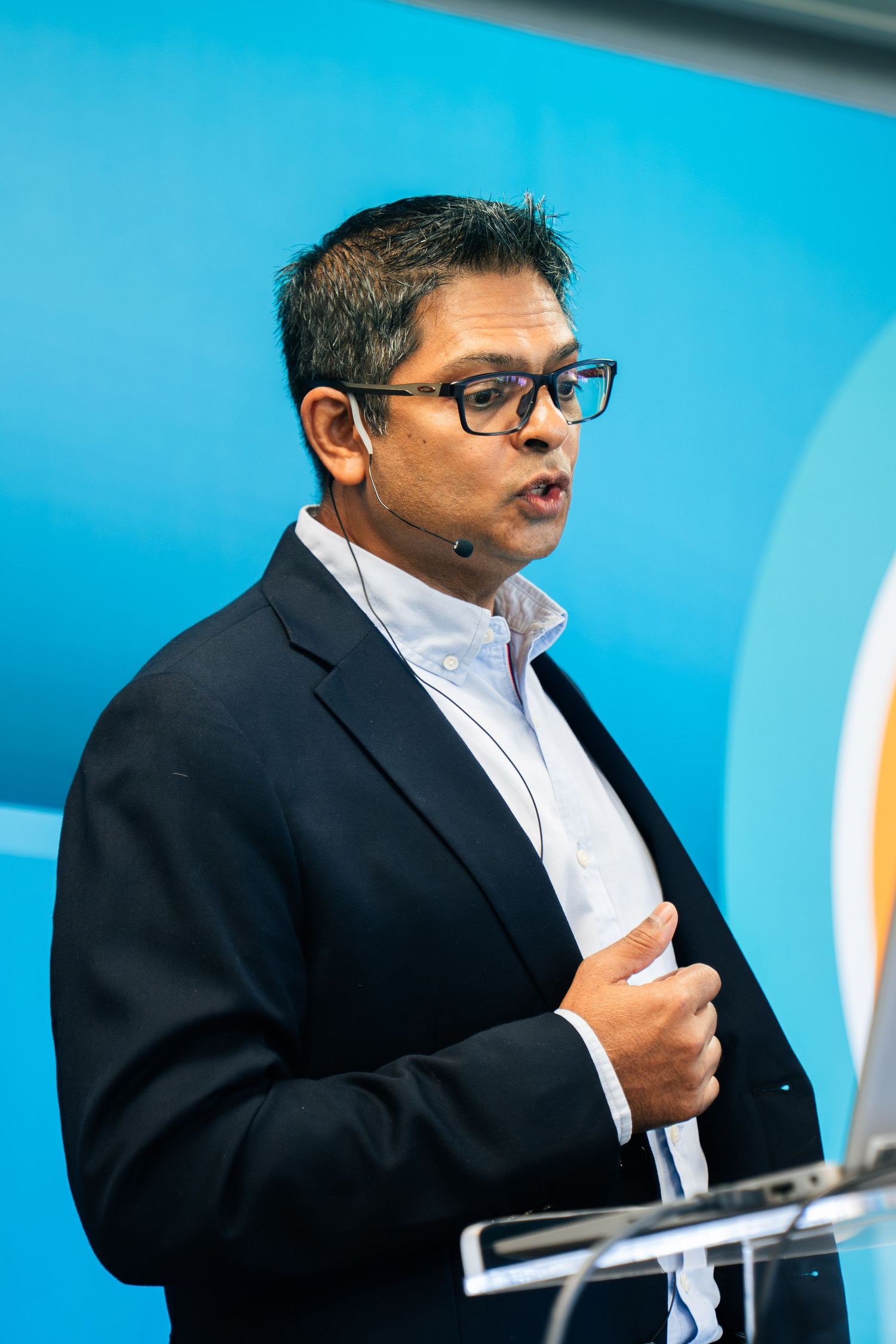 Professional male speaker wearing glasses presents at lectern with microphone in blue setting.