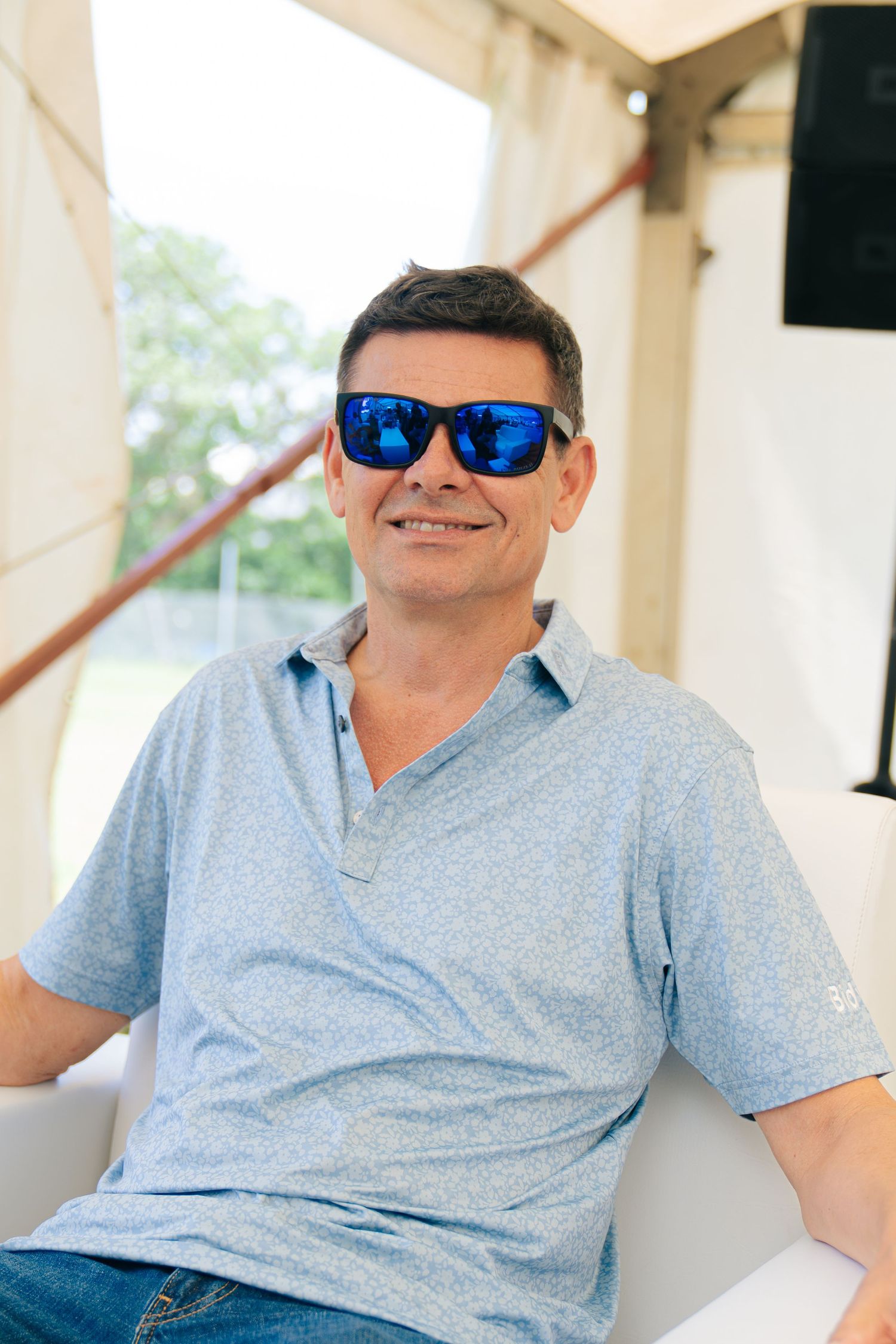Man smiling in blue mirrored sunglasses and light blue polo shirt seated outdoors at a sunny event.
