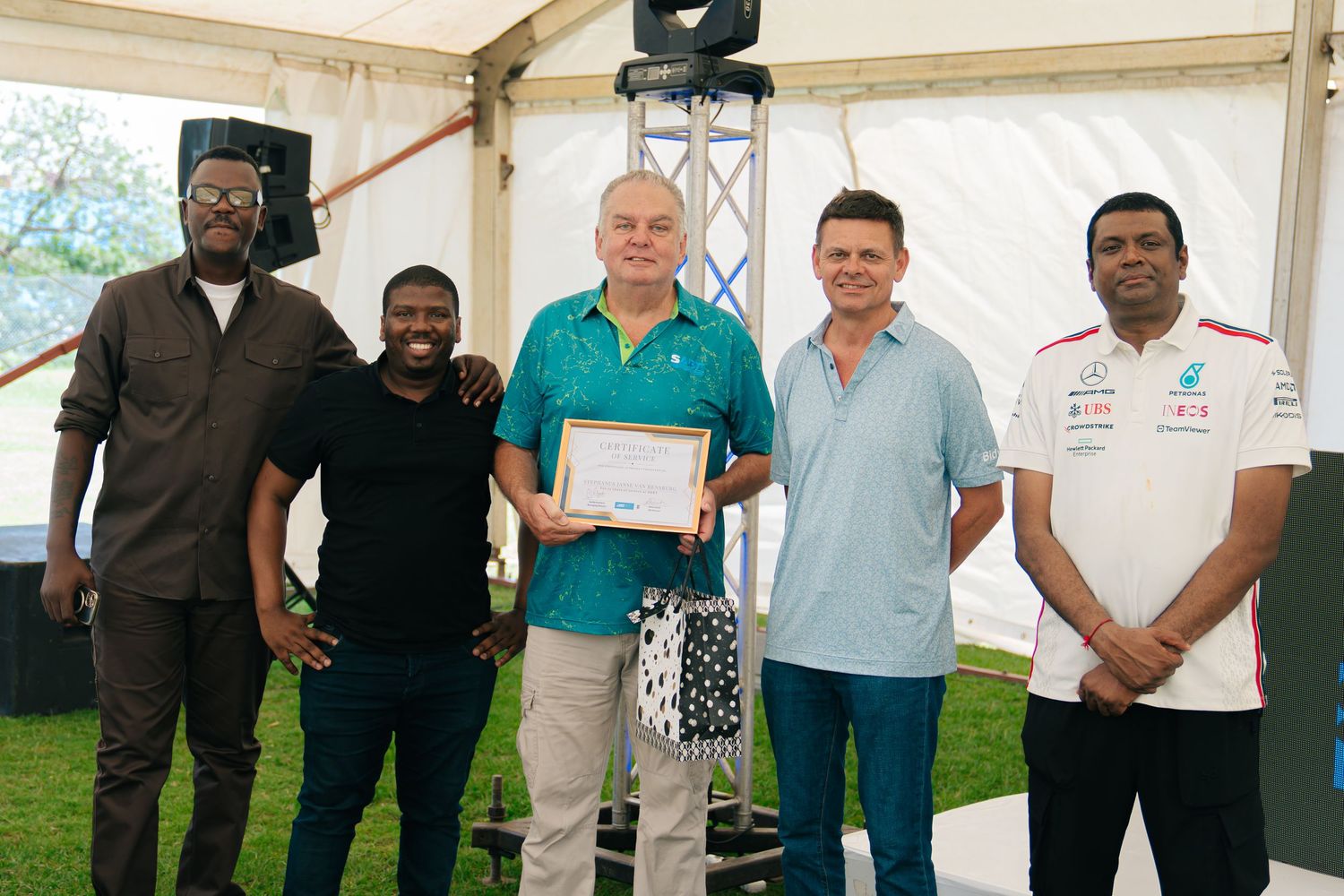 Five men posing together at an outdoor event, one holding a certificate of achievement under a white tent.