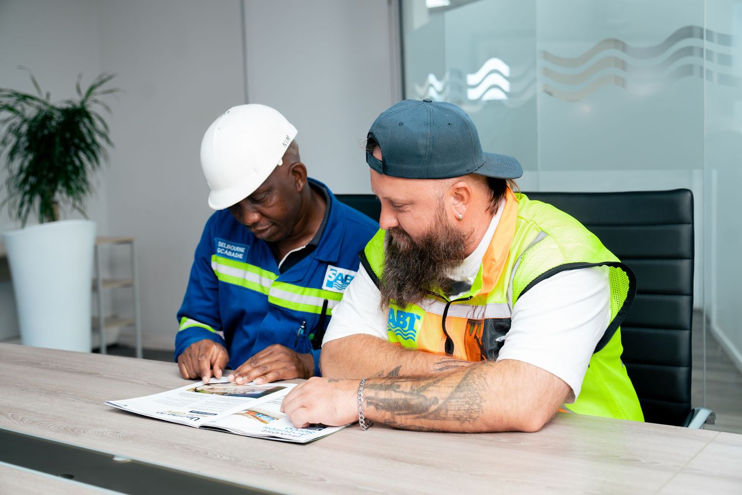 Two construction workers in safety gear review documents together at a conference table in a modern office.
