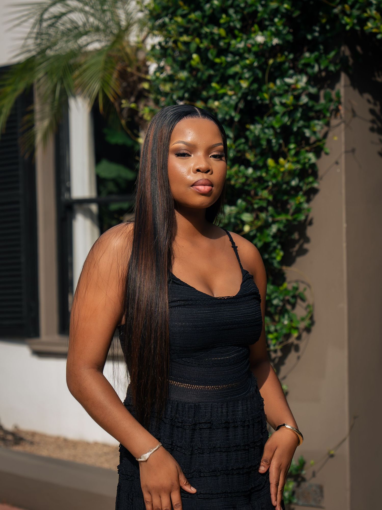 Young woman in black outfit with long straight hair poses outdoors near green hedges and palm trees.