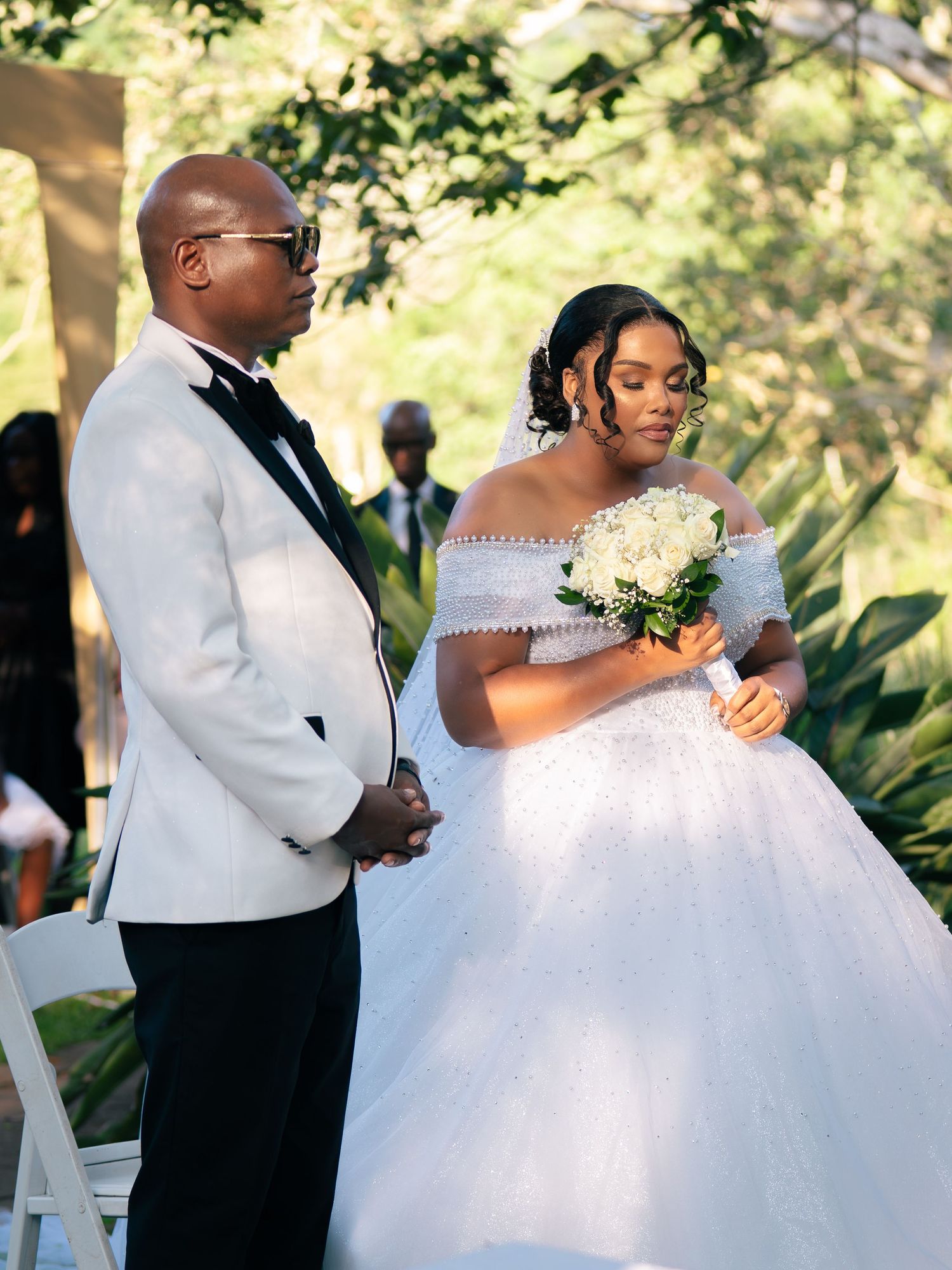 Bride in off-shoulder ball gown holding white bouquet stands with groom in white tuxedo at outdoor wedding ceremony.