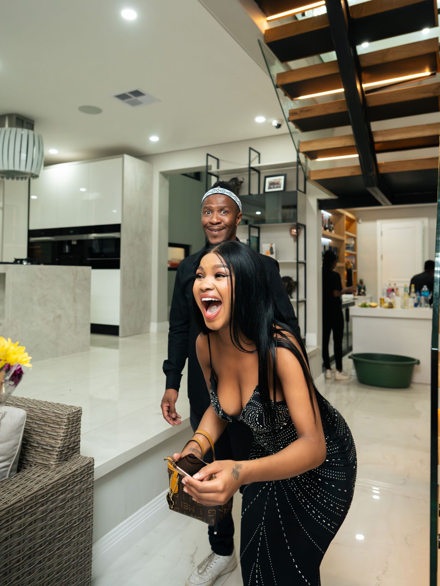A joyful woman in a black sequined dress laughs while a man smiles behind her in a modern luxury home interior.