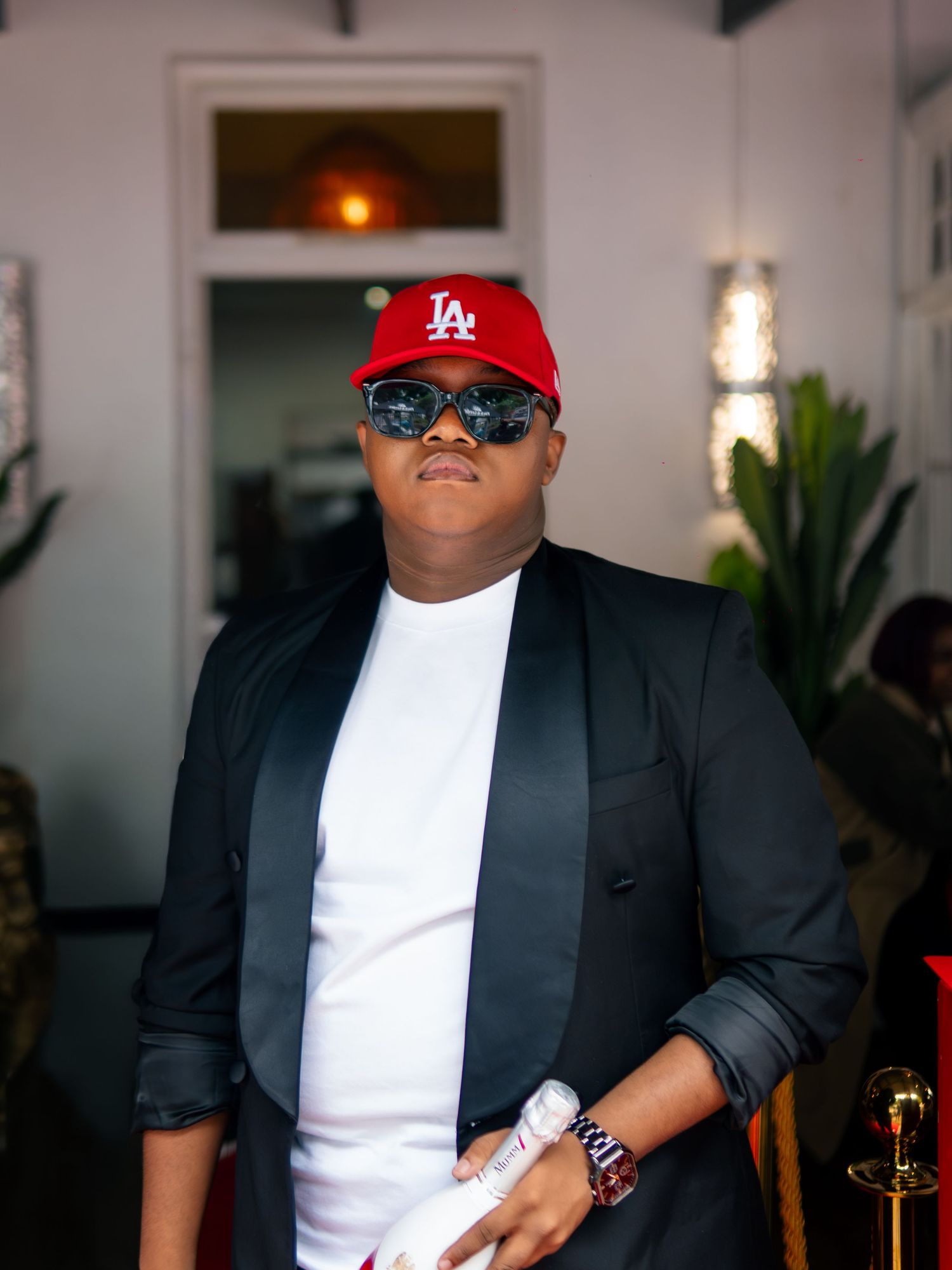 Stylish man wearing red LA cap, sunglasses, black blazer, and white t-shirt posing confidently indoors.