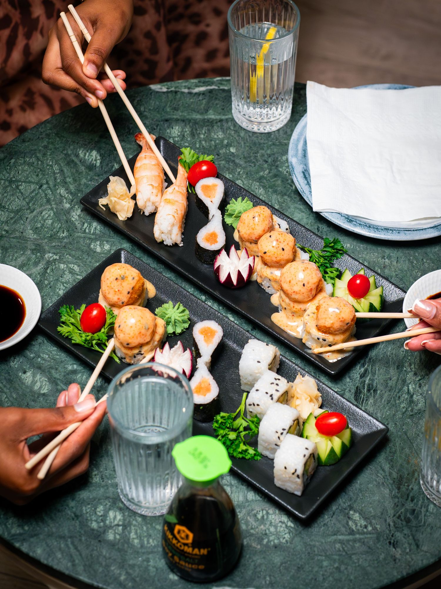 Two people sharing sushi platters with chopsticks on a green marble table with soy sauce and drinks.