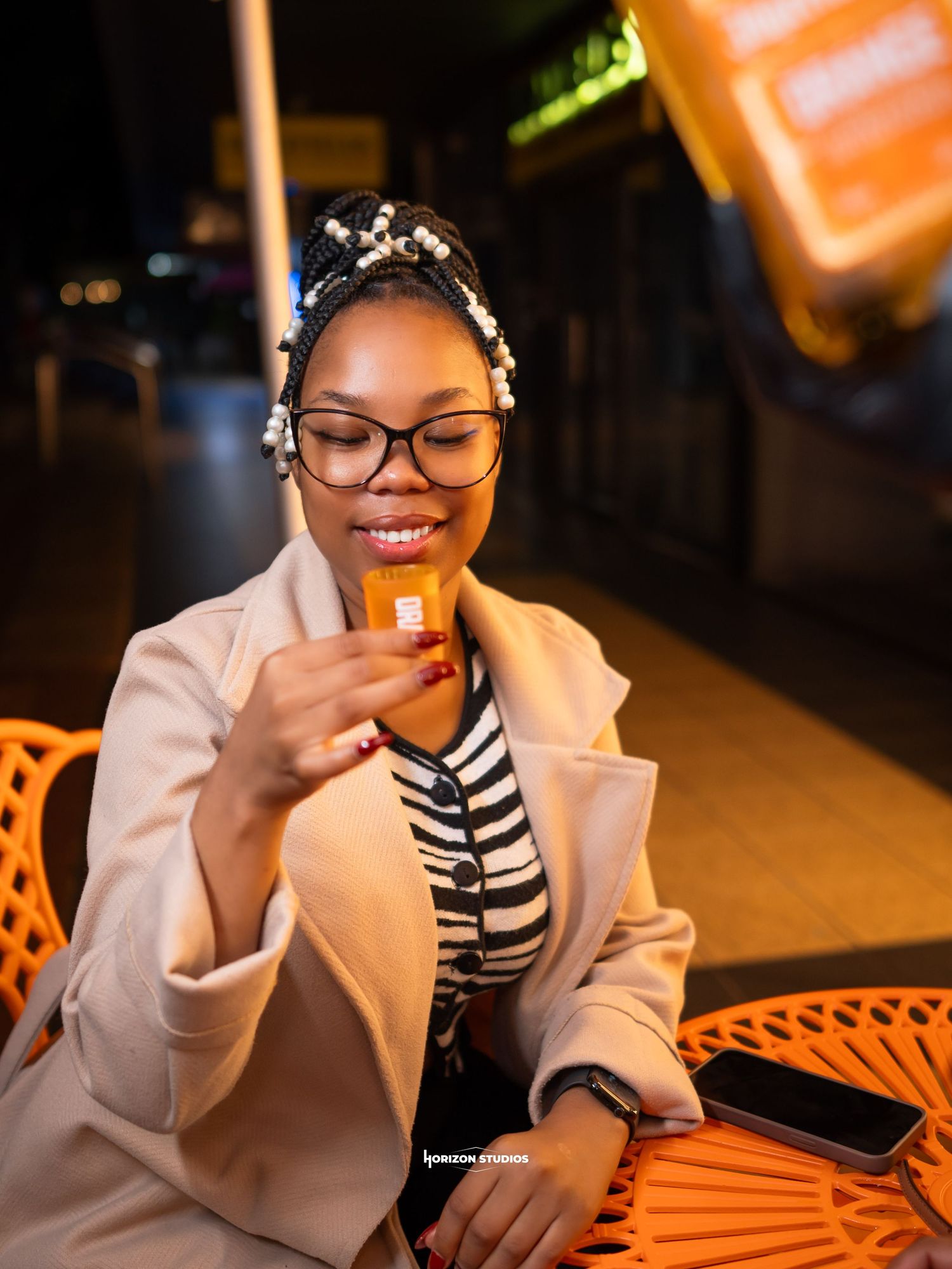 A young Black woman with braids and glasses smiles while holding a credit card at an outdoor cafe at night.