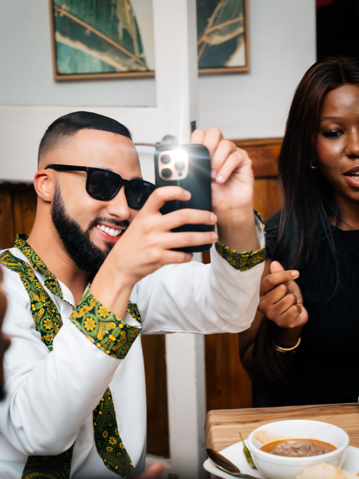 Man in sunglasses and African print shirt photographs food at restaurant table with woman beside him.