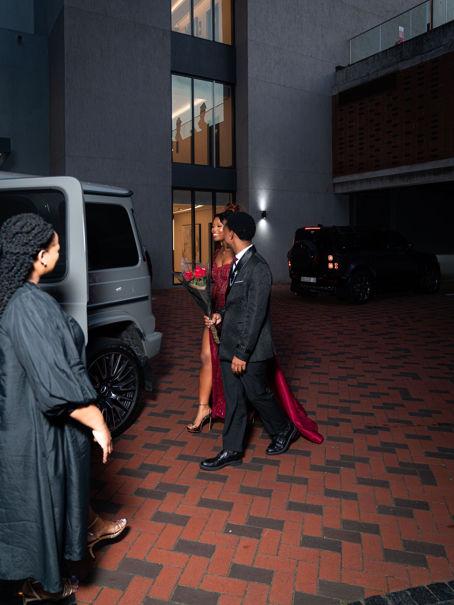 Elegant couple in formal attire walking toward building entrance at night, woman in red gown, man in suit.