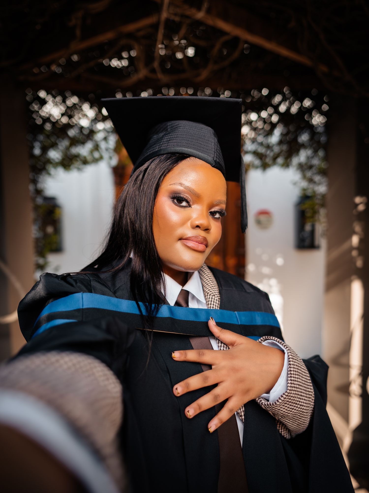 A young woman in graduation cap and gown with blue trim takes a selfie, smiling proudly at her academic achievement.