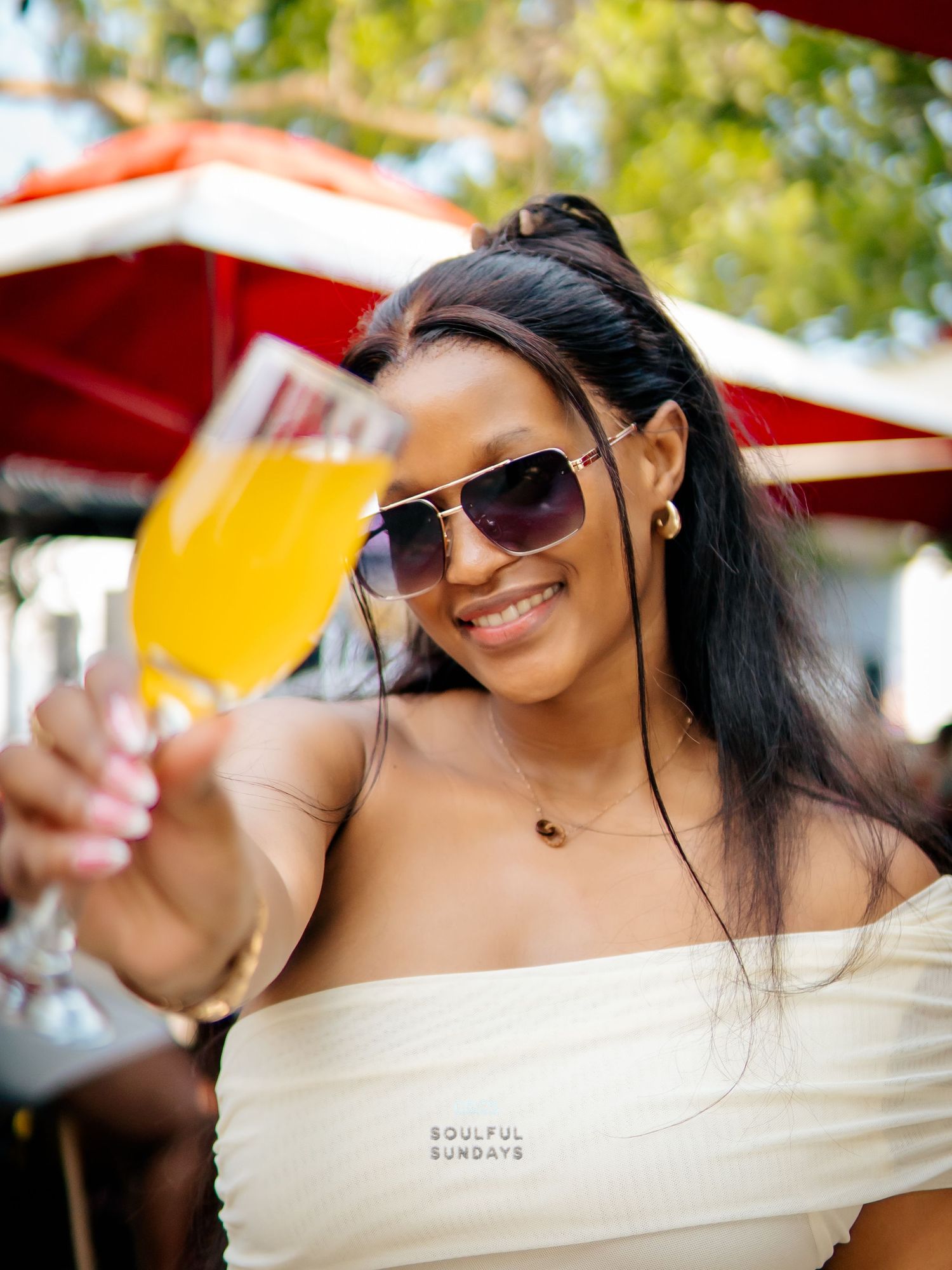 Woman in white off-shoulder top and aviator sunglasses toasting with a mimosa at an outdoor brunch event.