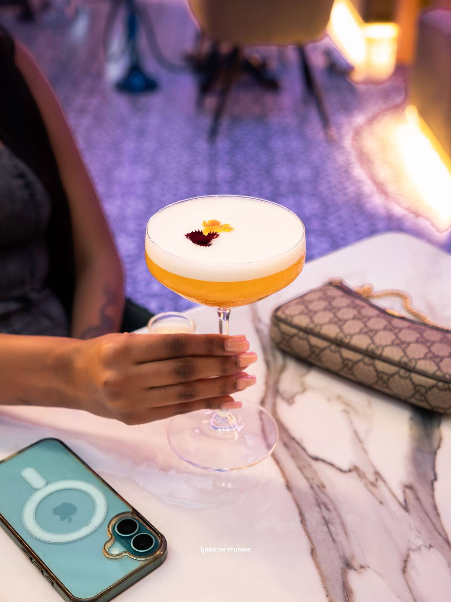 Hand holding a frothy yellow cocktail in a coupe glass on a marble table beside a Gucci bag and iPhone.