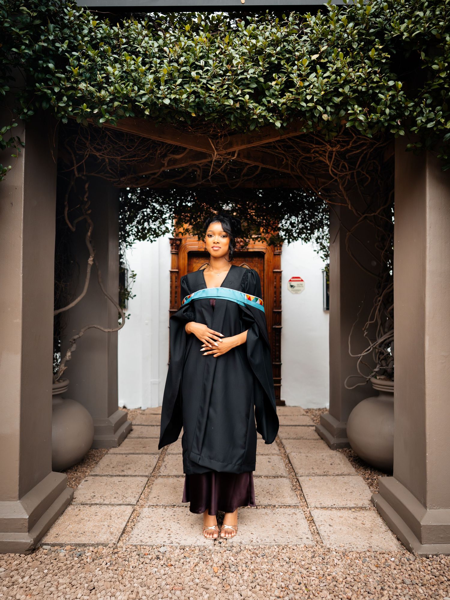 Woman in black graduation gown with blue sash stands between stone pillars beneath green foliage archway.