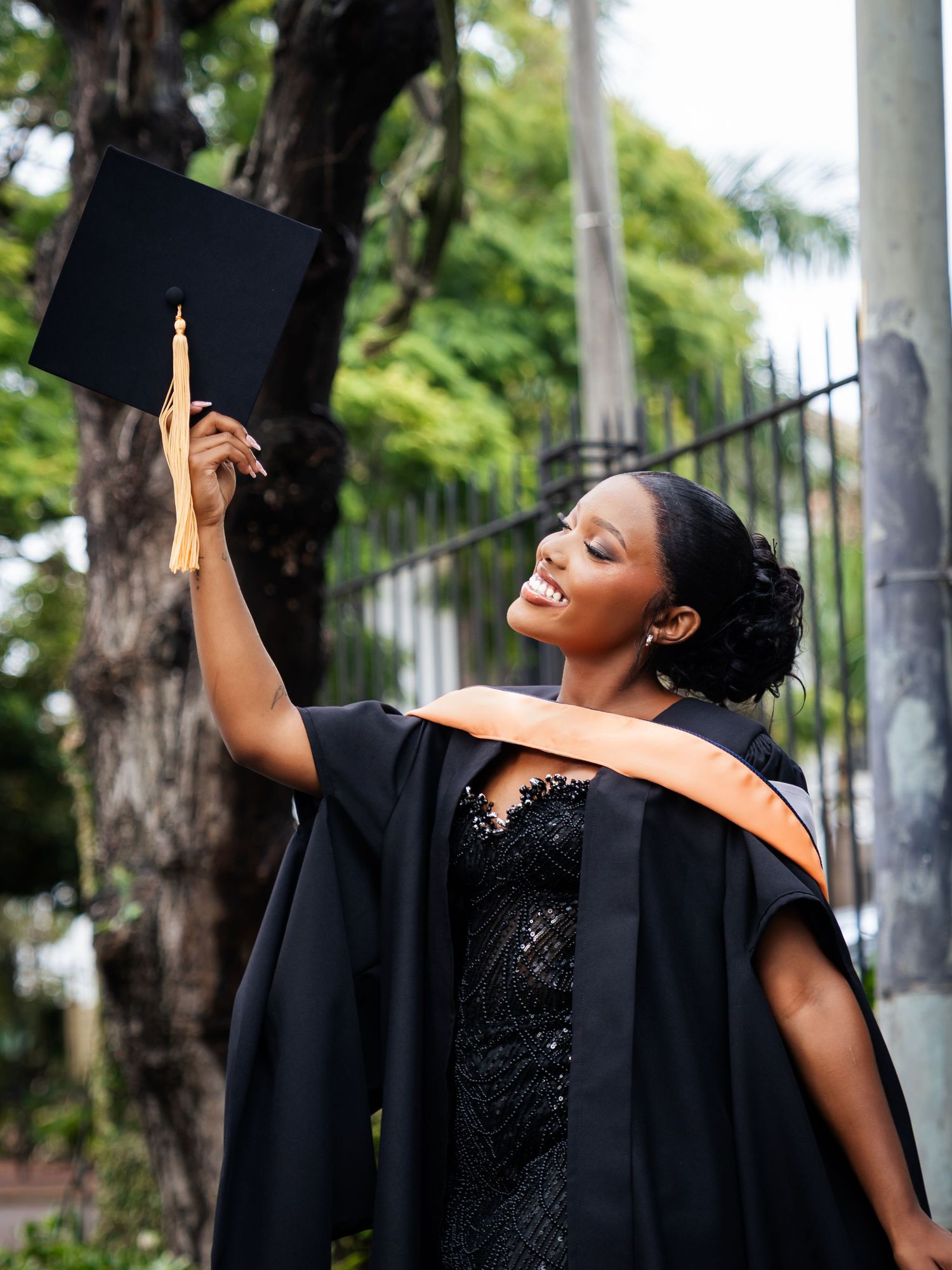 Smiling graduate in black gown with orange sash holding mortarboard cap aloft outdoors.