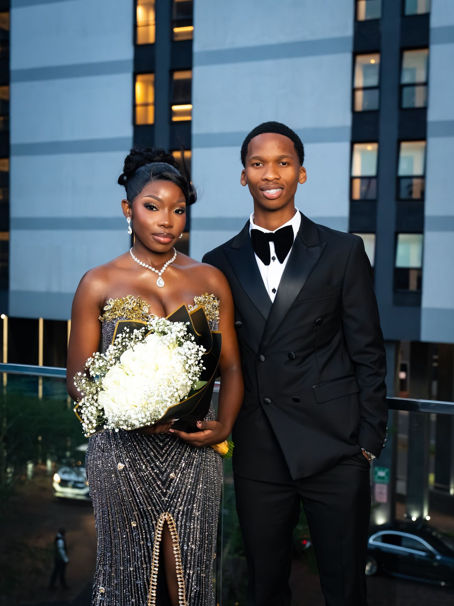 Elegant couple in formal attire, woman in beaded gown with bouquet, man in black tuxedo, posing outdoors at night.