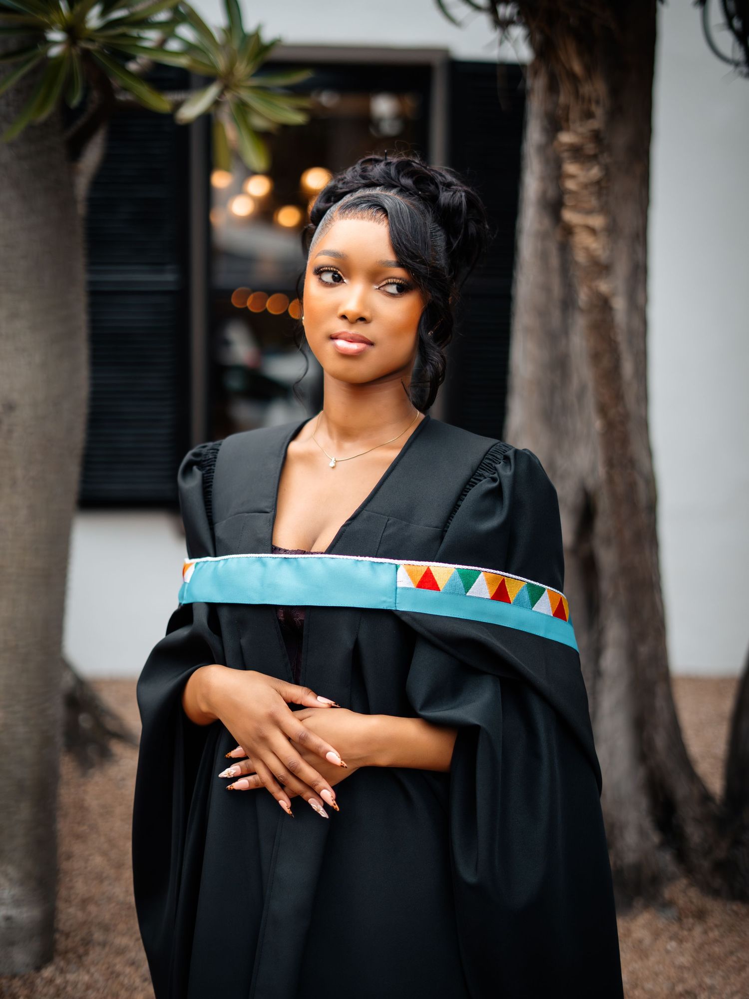 Young woman in black graduation gown with colorful African-inspired sash poses outdoors near trees.