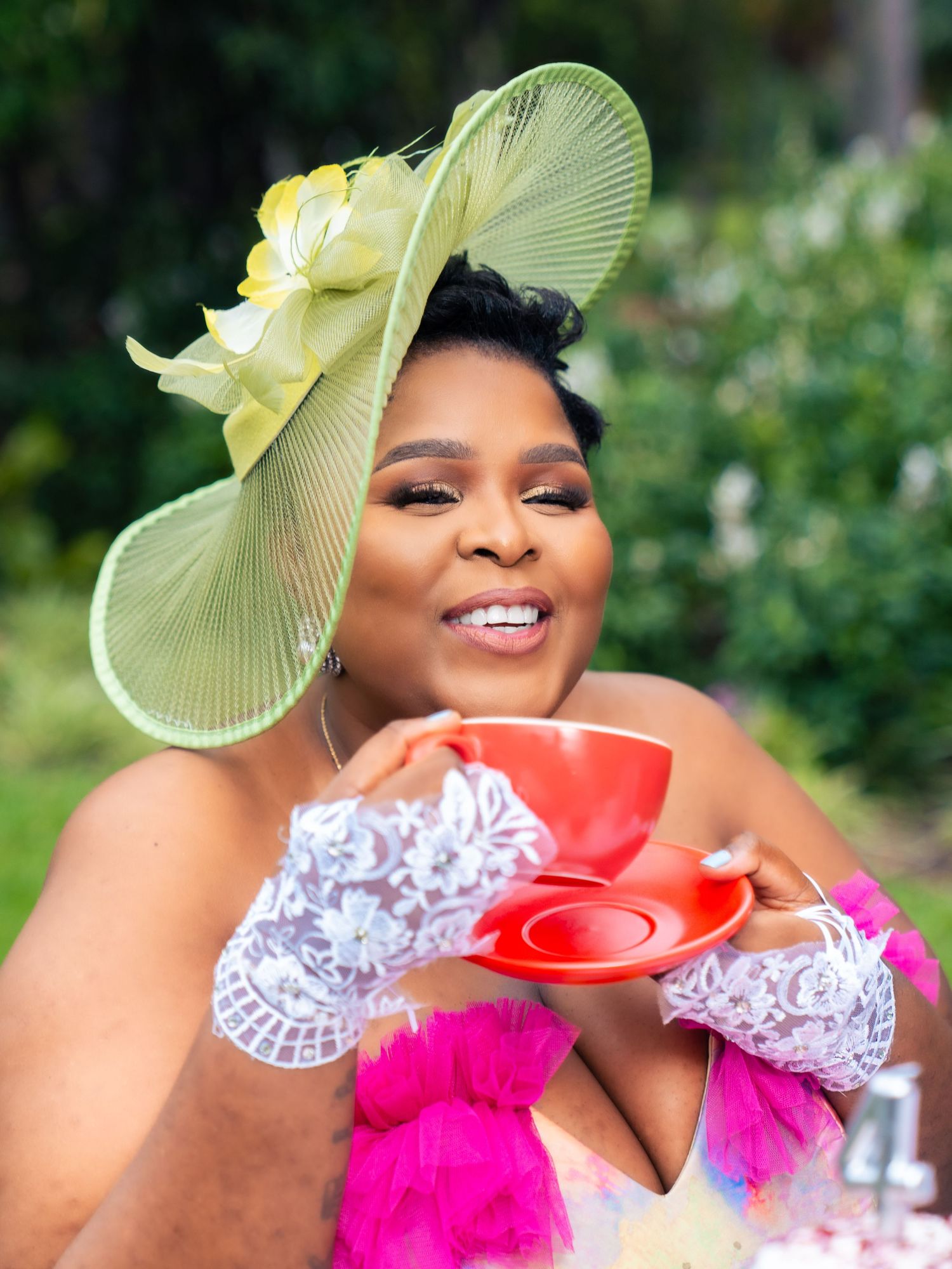 Woman in elegant green fascinator hat holding red teacup, wearing lace gloves and pink feather outfit at garden party.