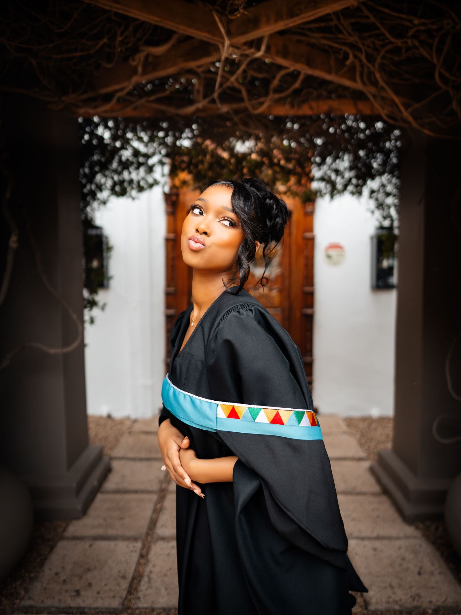Young woman in black graduation gown with colorful sash poses gracefully outdoors near elegant white building.