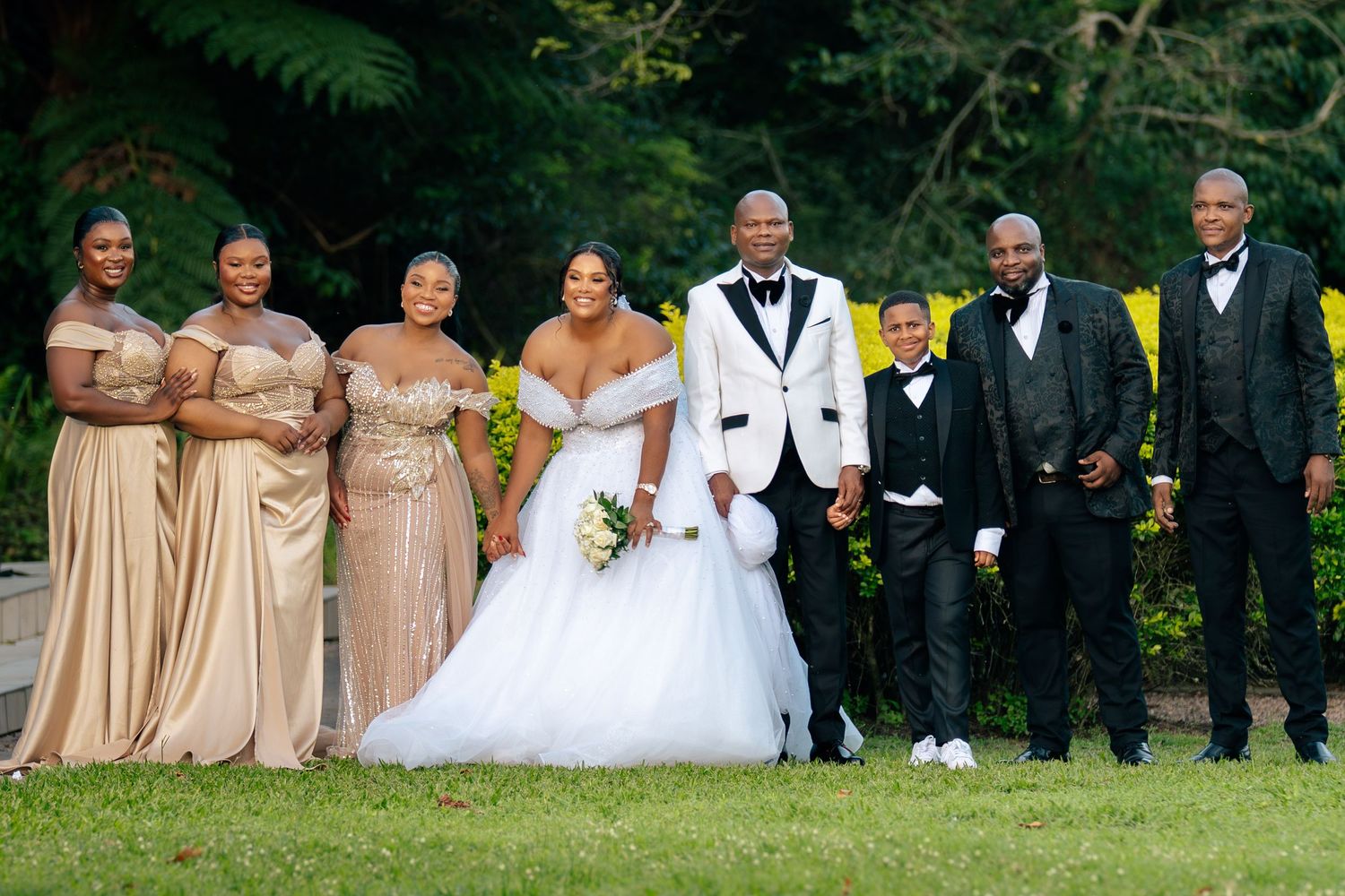 Wedding party posing outdoors, bride in white gown with groom in white tuxedo, flanked by bridesmaids in gold and groomsmen.