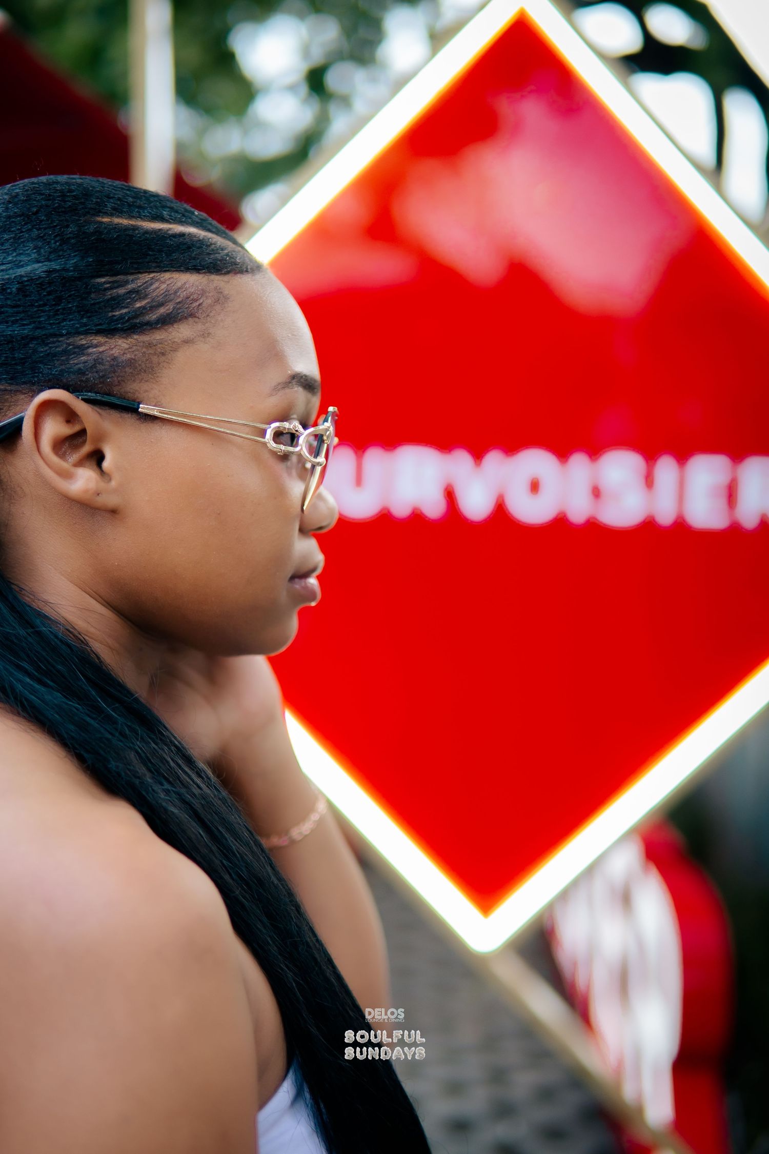 A young Black woman with braided hair and glasses poses in profile before a glowing red Courvoisier neon diamond sign.