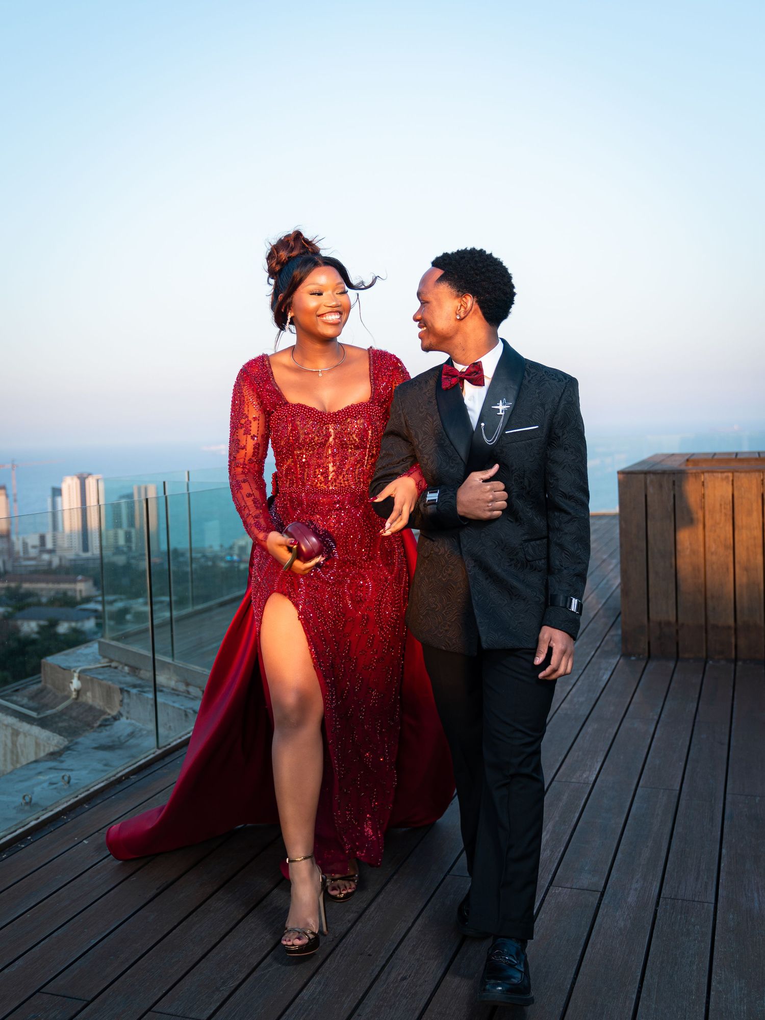 Elegant couple in formal attire on rooftop, woman in red lace gown, man in black tuxedo with bow tie.
