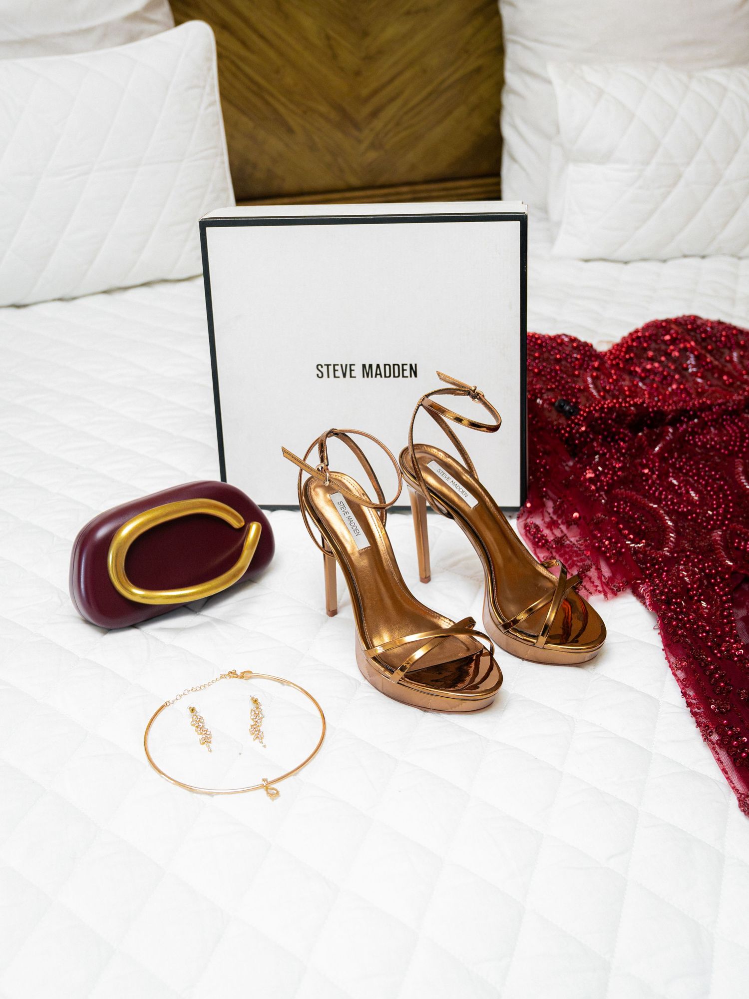 Gold Steve Madden strappy heels displayed on white bed with red sequin dress, burgundy clutch, and gold jewelry.