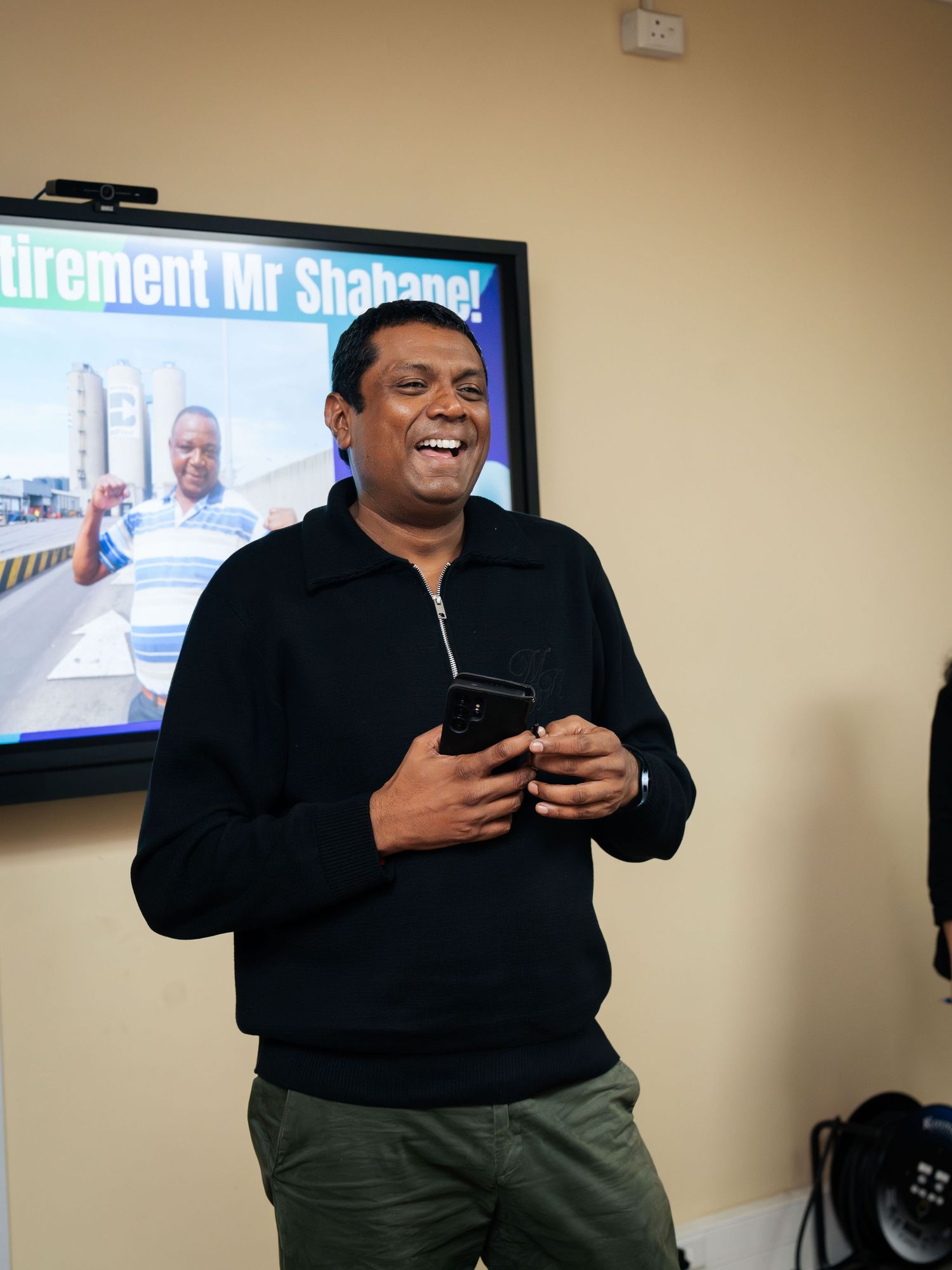 Man in black sweater smiling and holding phone at Mr. Shabane retirement party with TV screen in background.
