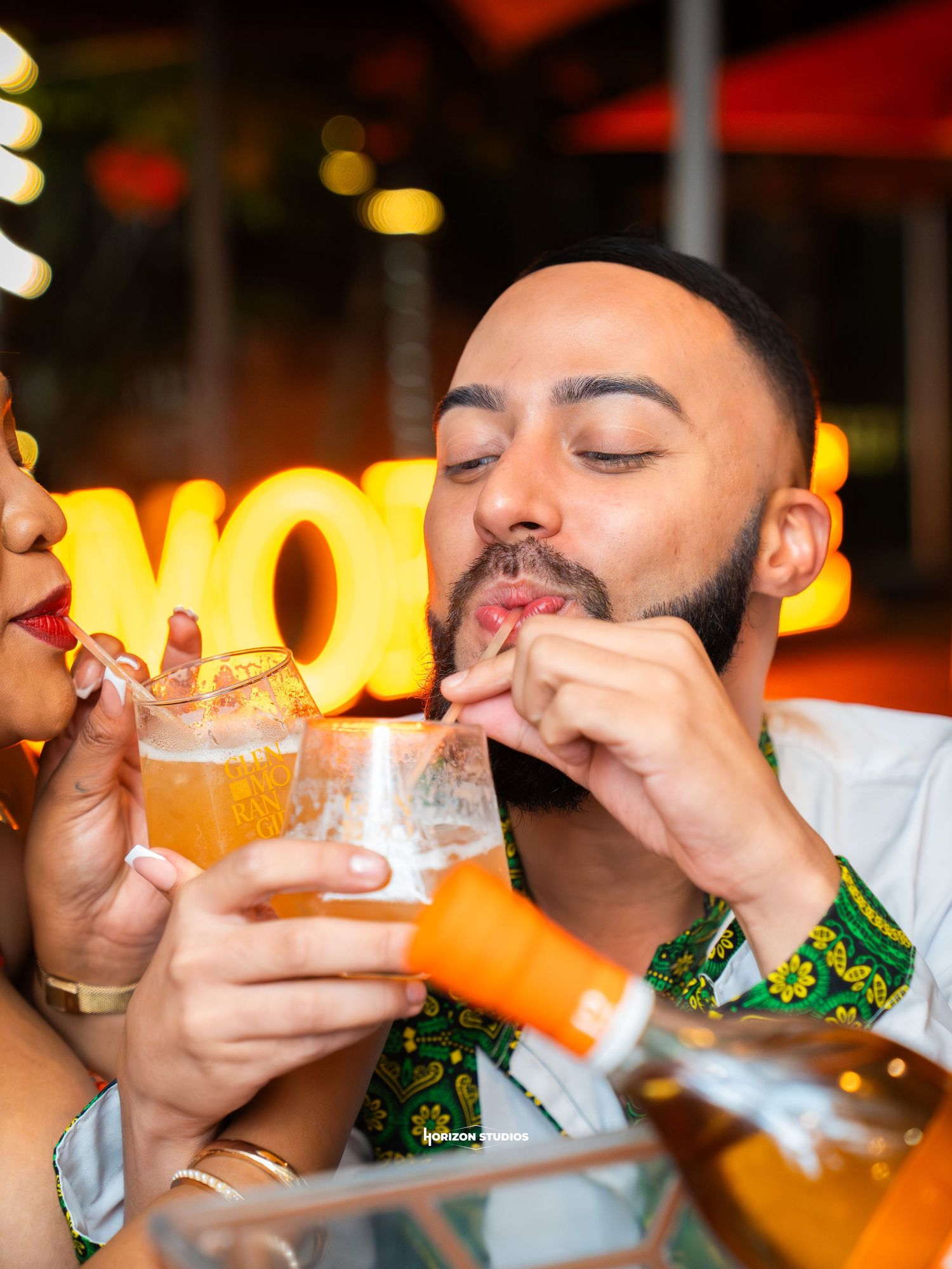 Man in green floral shirt sipping a cocktail through a straw at a vibrant bar with neon lighting.