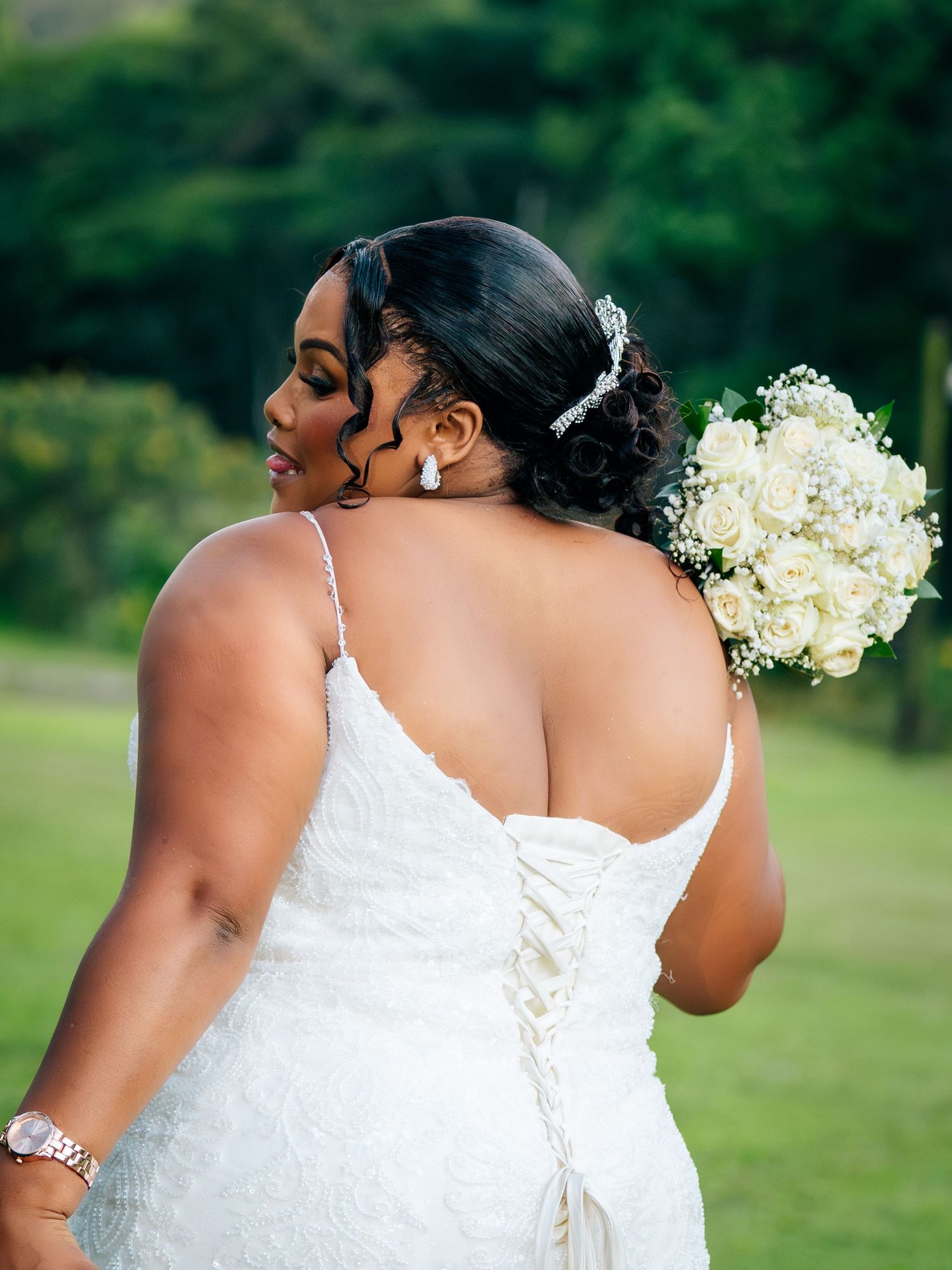 Bride in white lace gown holding white rose bouquet, showing elegant back detail with corset lacing outdoors.