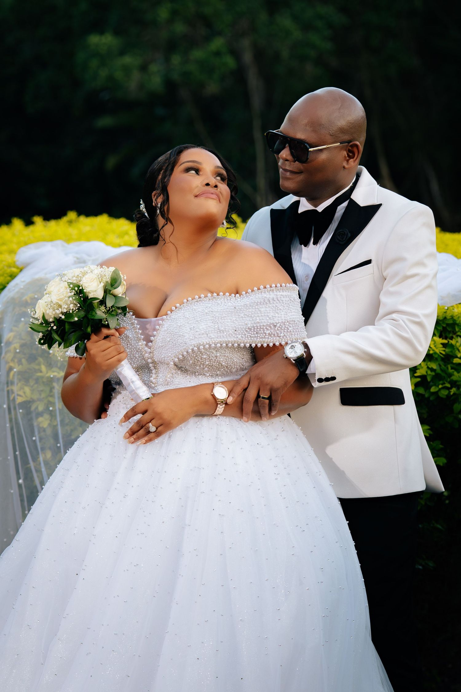 A bride in a white off-shoulder ball gown holds flowers as a groom in a white tuxedo embraces her outdoors.