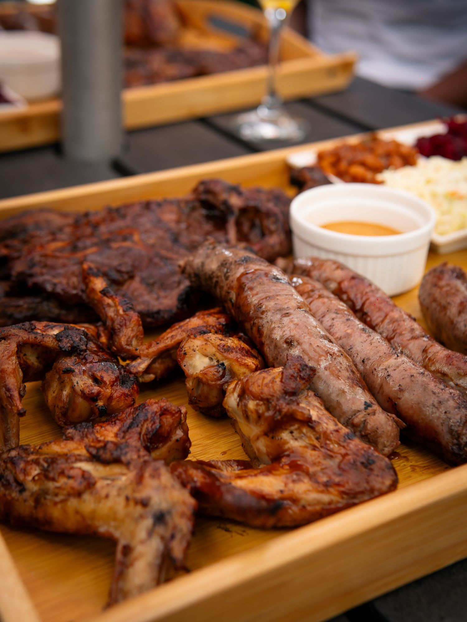 A wooden platter of grilled meats including chicken wings, sausages, and ribs served with dipping sauce.