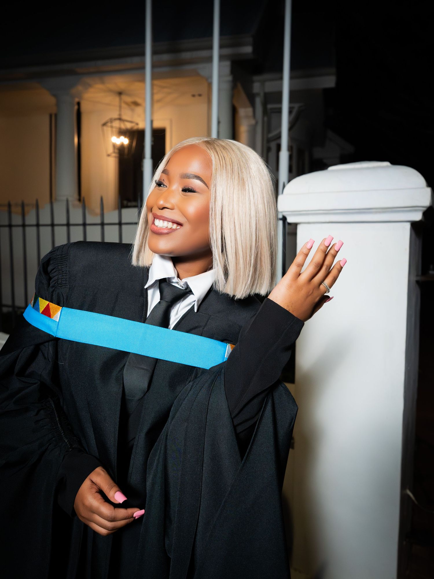Smiling graduate in black gown with blue hood posing against white pillar at night.