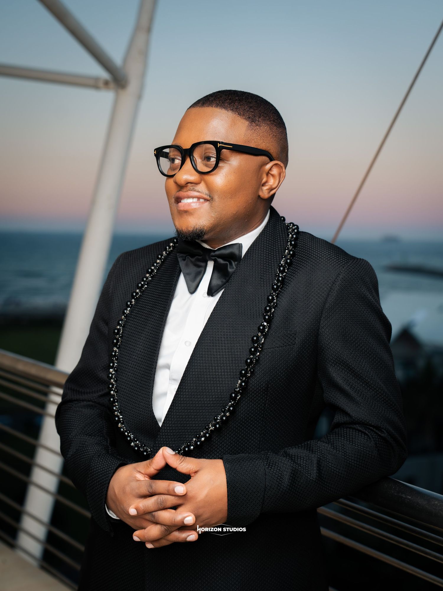 Young man in black tuxedo and bow tie with beaded necklace smiling outdoors at sunset near ocean.