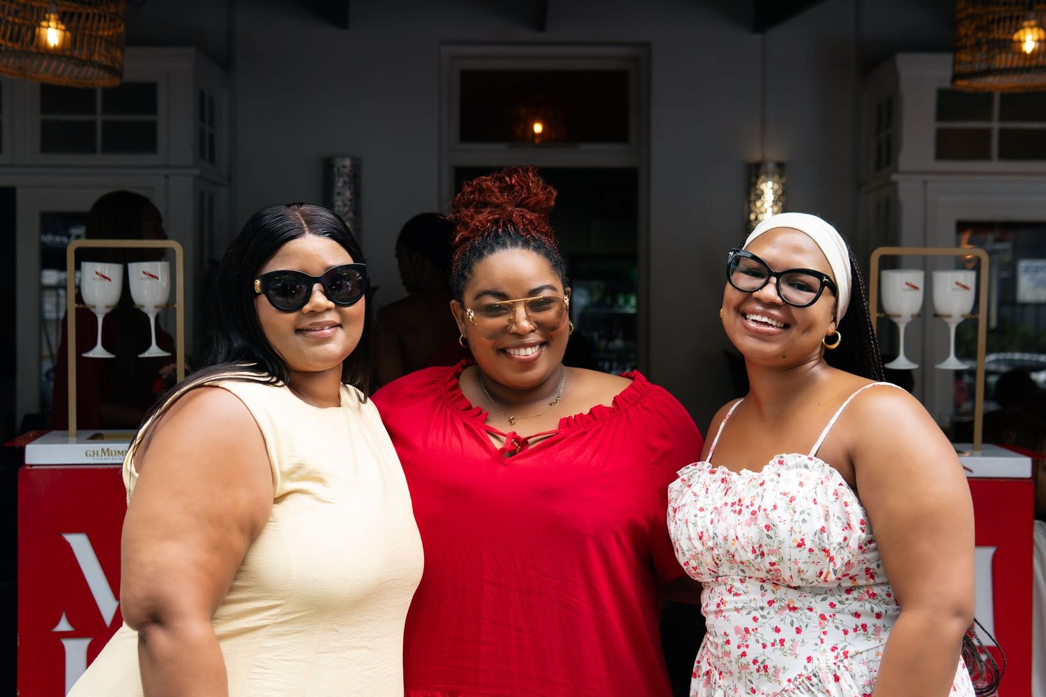 Three women smiling outdoors at an event, wearing sunglasses, dressed in red, white, and floral outfits.