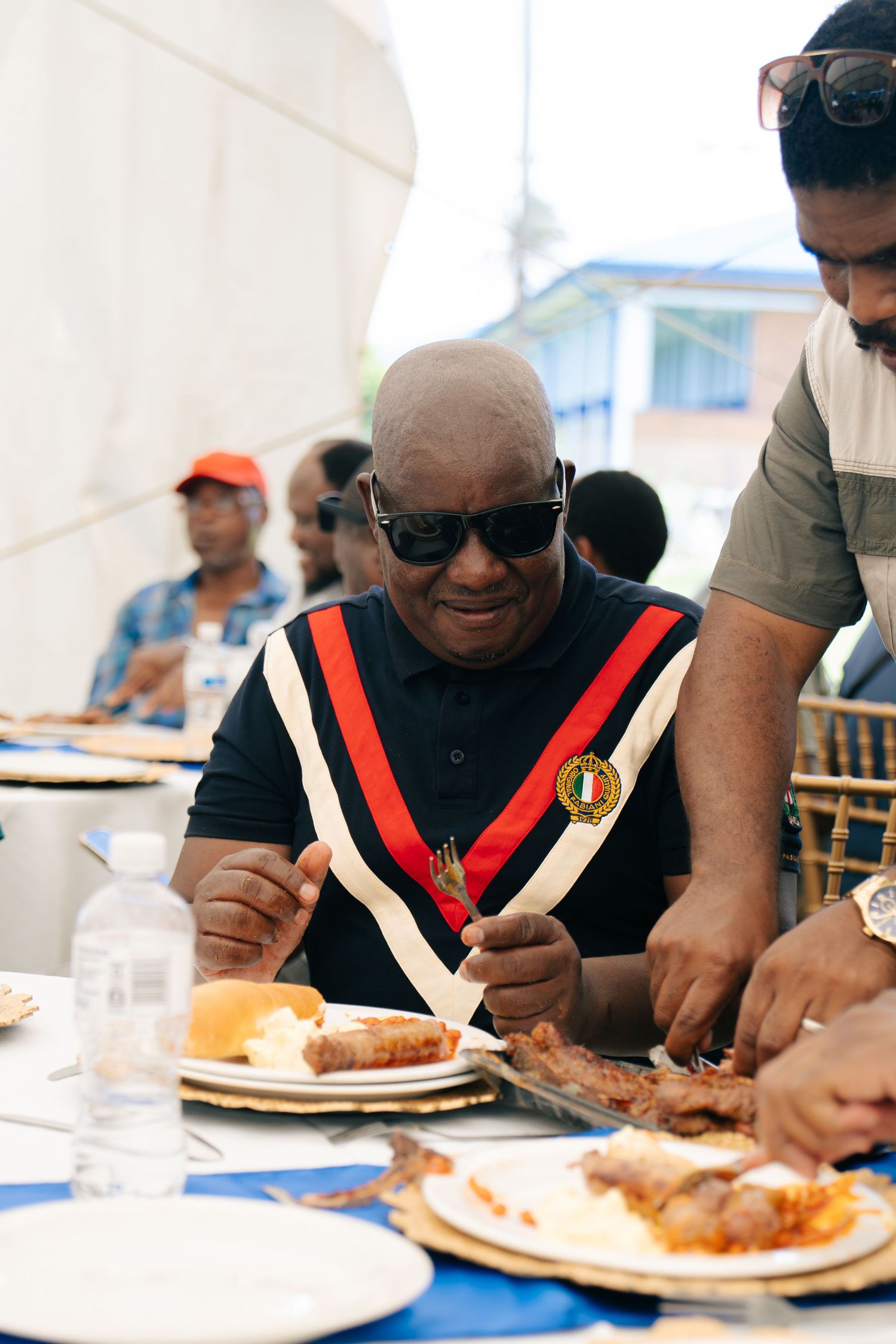 Bald man wearing sunglasses and a medal sash enjoys a meal at an outdoor event with other guests.