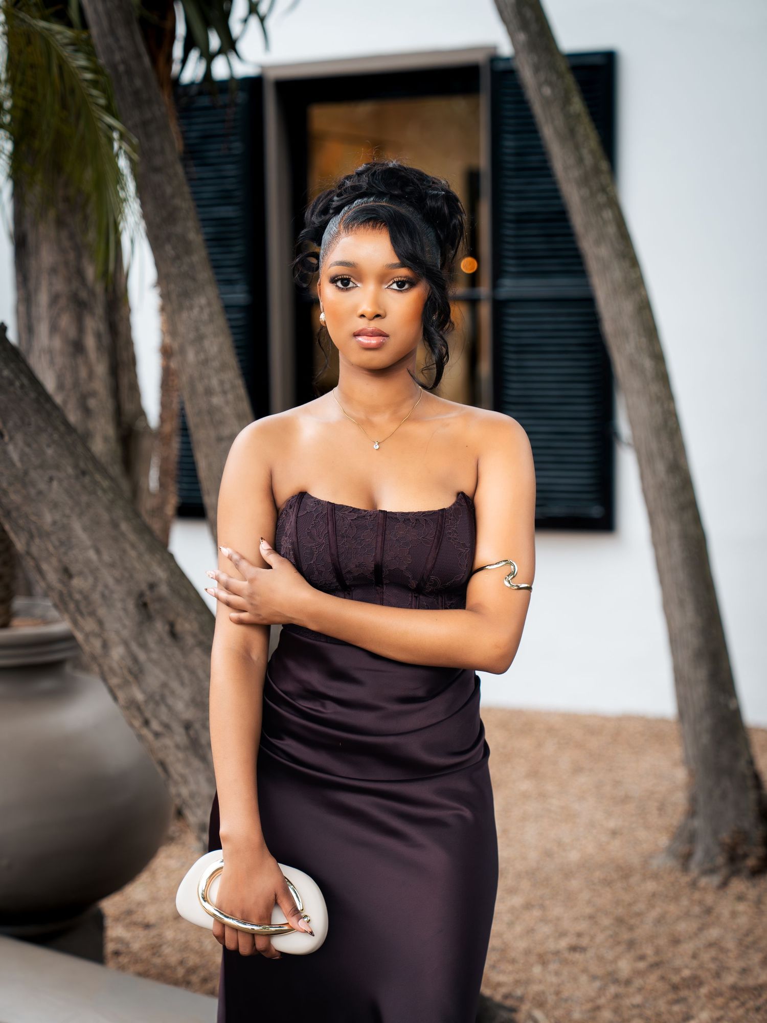 Elegant woman in strapless dark purple gown holding white clutch purse outdoors near palm trees.