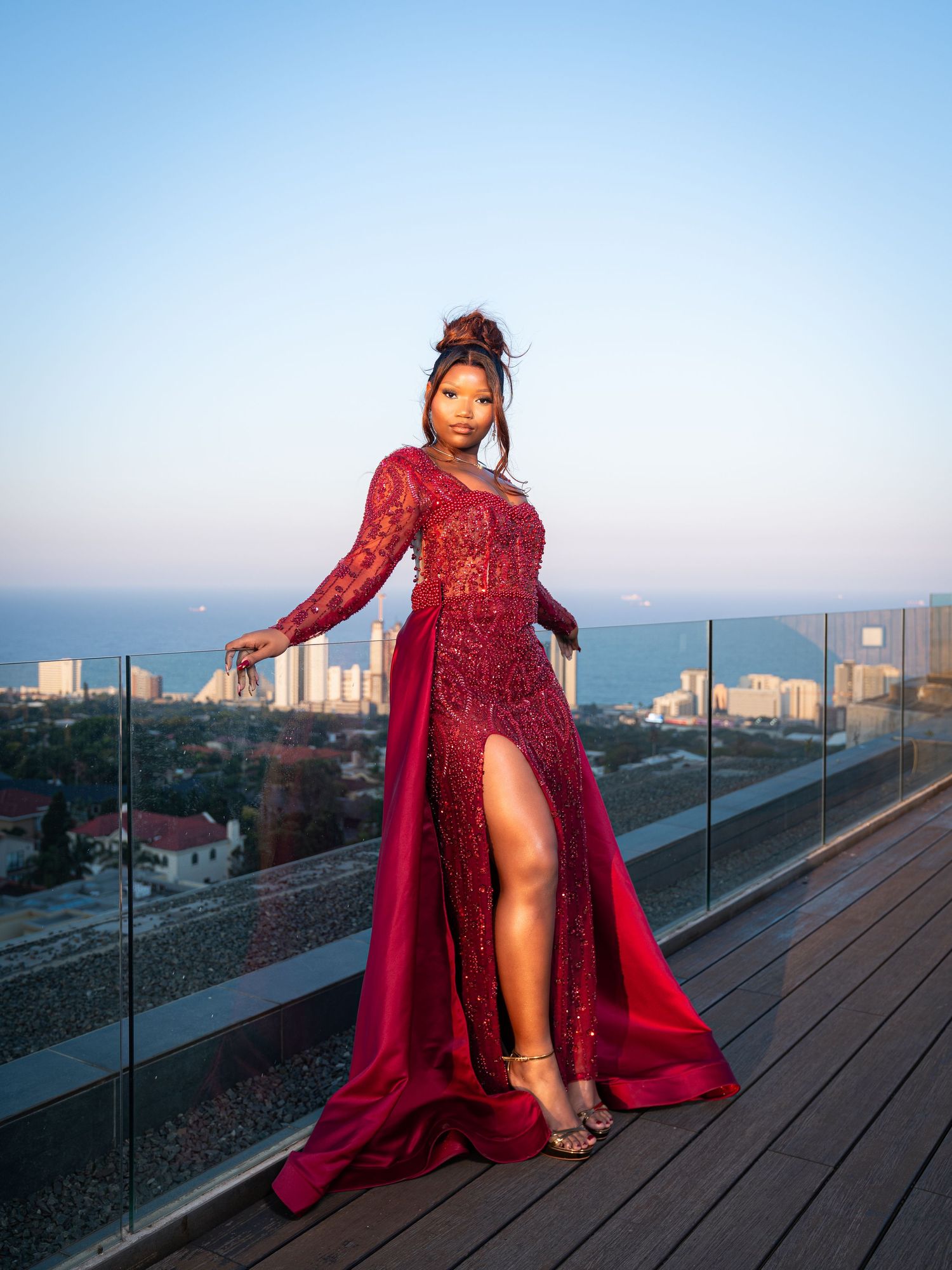 Woman in elegant red lace gown with slit poses on rooftop terrace overlooking coastal city skyline at dusk.