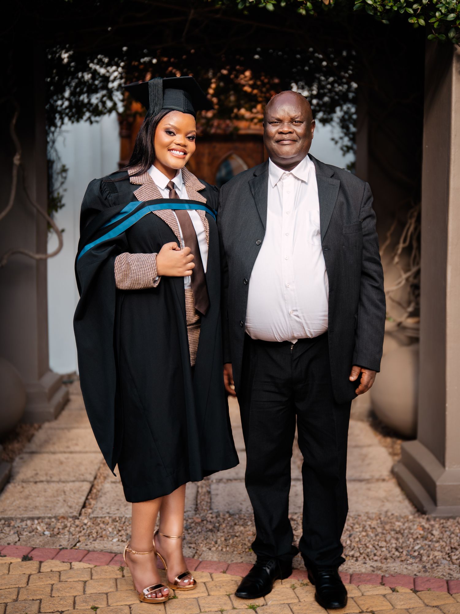 Graduate in black academic gown and cap poses proudly with a man in a suit at an outdoor graduation ceremony.